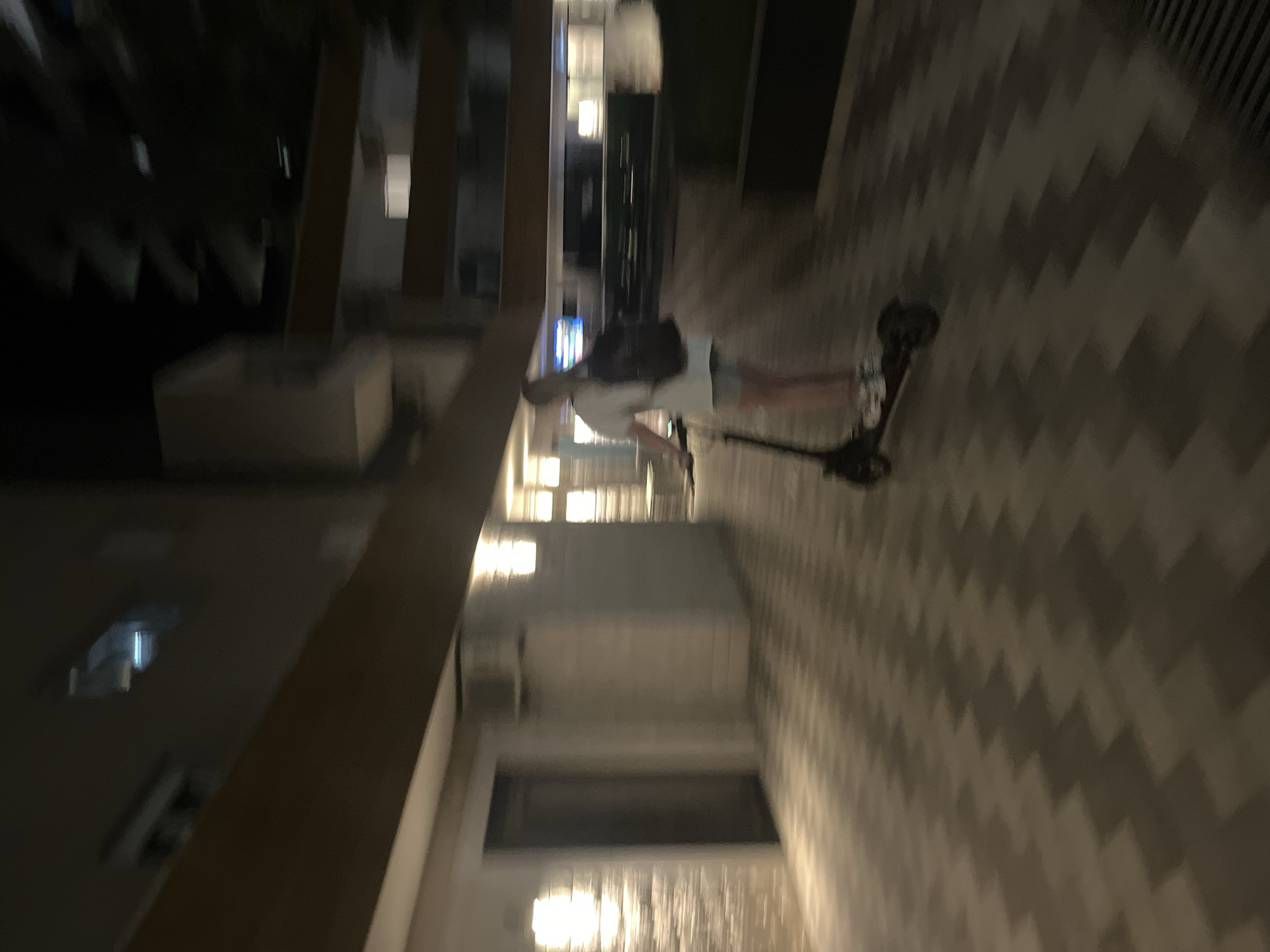 A person rides an electric scooter at night on a zigzag patterned walkway outside of a building. The person is wearing a white t-shirt, grey shorts, and a black backpack. The walkway is illuminated by lights on the building. The image appears to be taken in Budva, Montenegro, a city known for its historic old town and beaches.