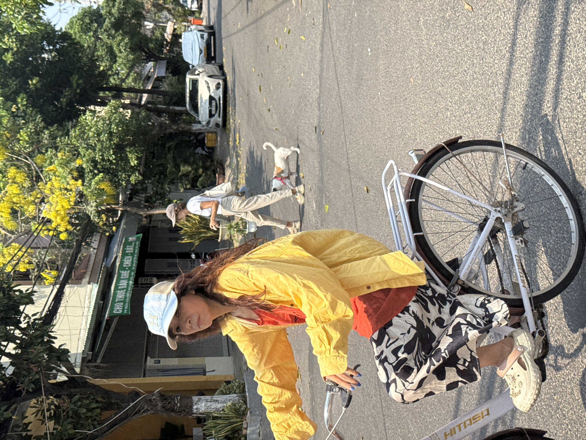 A woman is riding a bicycle on a street. She is wearing a yellow jacket, a red top, and patterned pants. She also has a baseball cap on her head. In the background, a couple is walking a dog. There are also cars parked on the street, as well as trees and buildings. The sign "CHO THUE BAN GHE - CHEN - BAT…" indicates the location might be in Đà Nẵng, Vietnam.