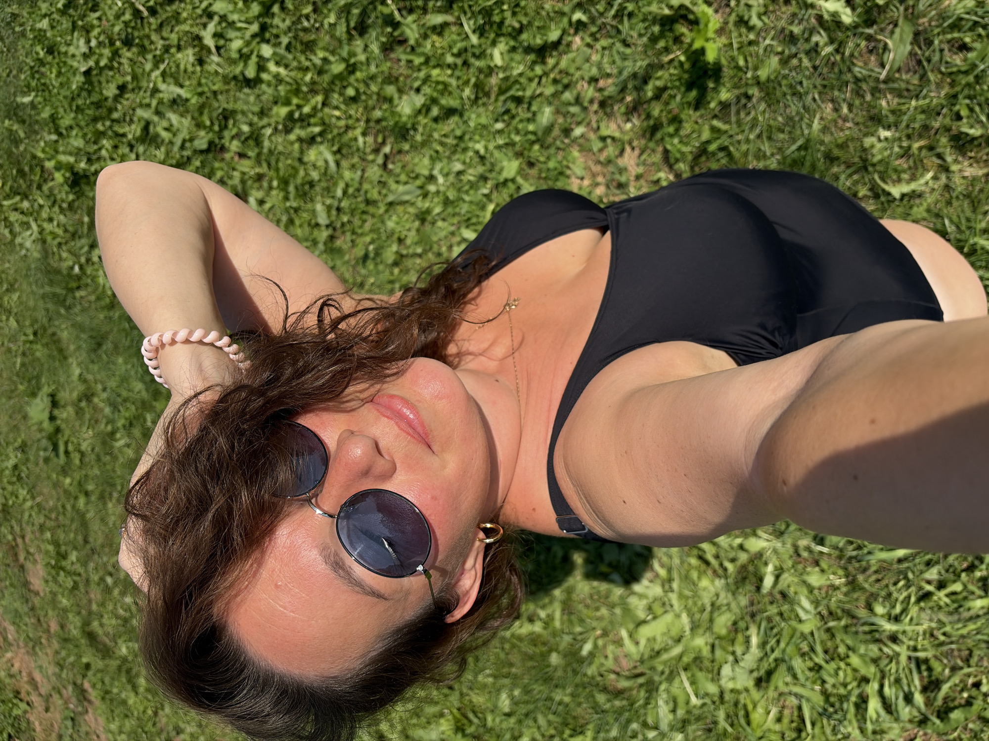 A woman is taking a selfie outdoors, with a grassy area visible in the background. She is wearing a black swimsuit and sunglasses, with her right arm raised behind her head. She has on a bracelet and hoop earrings.
