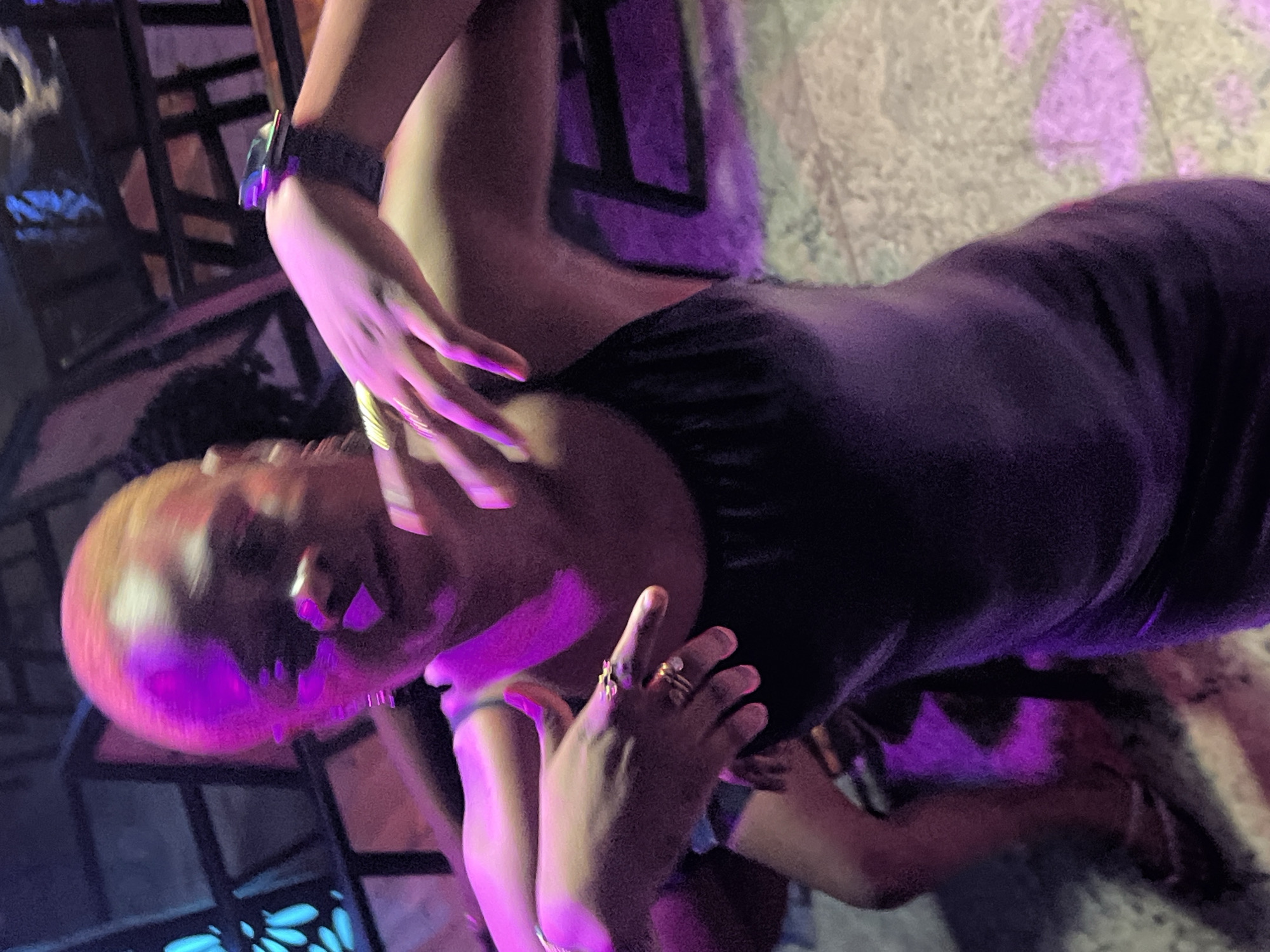 A person with short, dark hair is visible in the frame. They are wearing a dark top, and rings are visible on the fingers of one hand. The scene is illuminated with a purple light, and the background appears to be a mix of furniture and a patterned floor. No location is determinable.