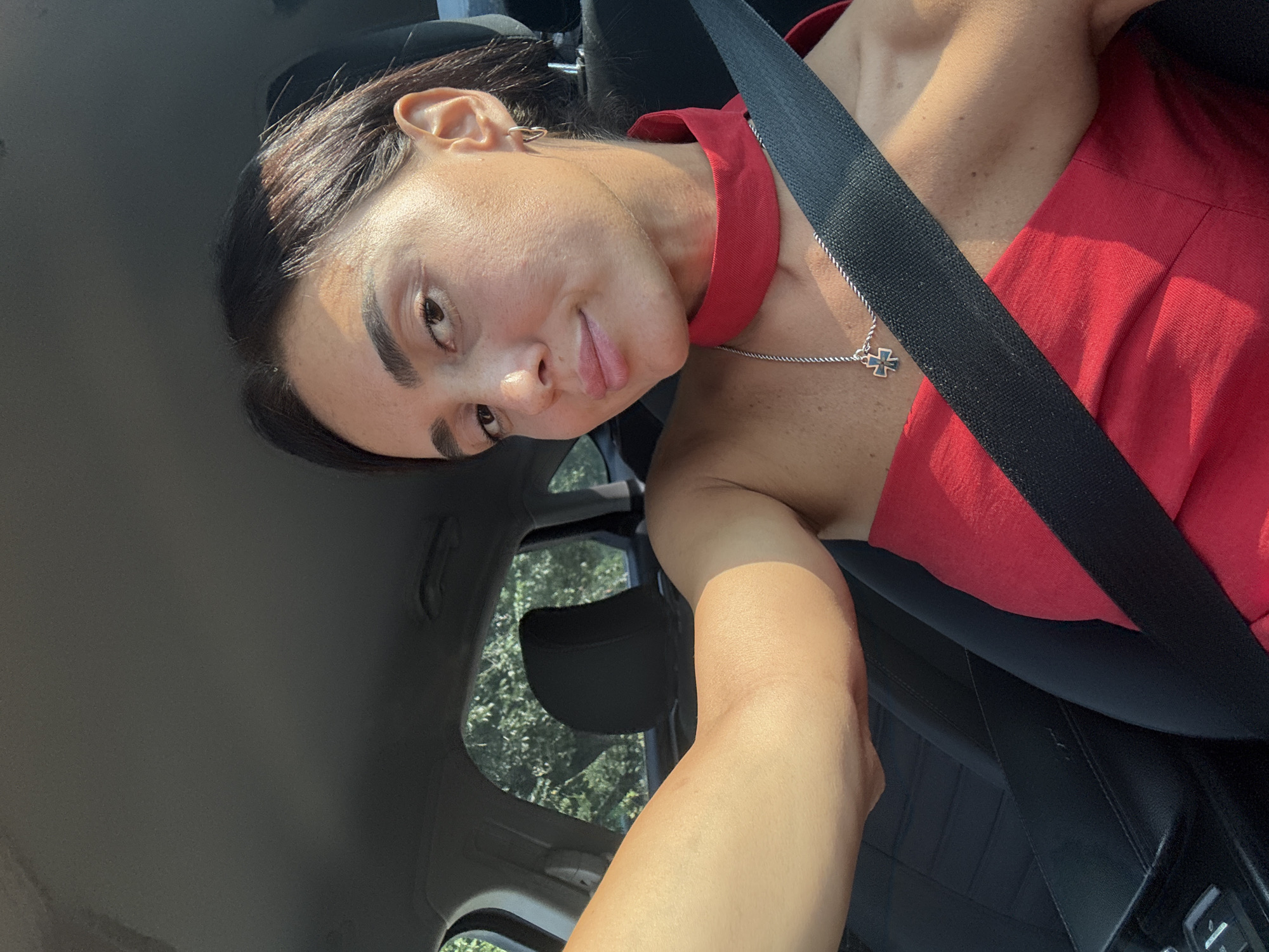 In this selfie taken from the driver's seat of a car, a woman with dark hair and light skin is looking at the camera with a slight smile. She is wearing a red top with a scarf-like neckline and a silver necklace with a cross pendant. Her left arm is visible, extended towards the camera. A black seatbelt is worn across her chest, and the interior of the car, including a headrest and part of the dashboard, is visible. Outside the car windows, green foliage is apparent, suggesting a setting with trees. The image appears to be taken during the daytime, with sunlight illuminating the scene. No specific location within Tbilisi, Georgia, can be determined from the image.