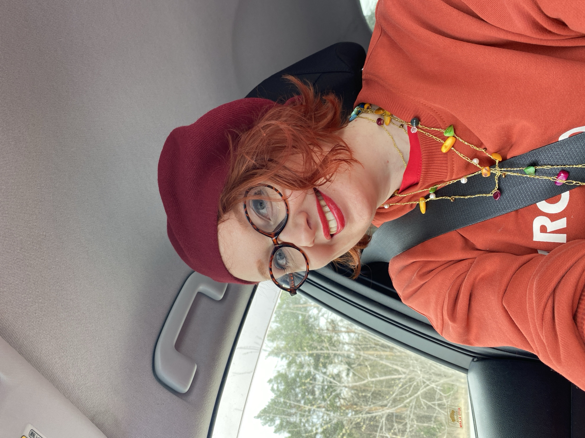 The image features a cheerful woman with reddish-orange curly hair and blue eyes, smiling broadly while seated in a car. She is wearing round, tortoise-shell frame glasses, bright red lipstick, and a dark red/maroon knit cap or beanie. Her attire includes an orange-red long-sleeved top, possibly a sweatshirt, which has some white text on the front, partially showing an 'R'. Adorning her neck are multiple necklaces with gold-colored chains strung with various colorful beads and small pearls. She is wearing a seatbelt across her chest.
The setting is the interior of a vehicle, with a light grey headliner visible above and a grey grab handle to her right. Through the car window to her left, bare trees under what appears to be an overcast sky are visible, suggesting a cooler season such as late fall, winter, or early spring. The scene indicates daytime. On the car window, there's a sticker that, when oriented correctly, appears to have the word "TOYOTA" (upside down in the image) and other indistinguishable text. The woman's wide smile conveys a sense of happiness or contentment. This scene is set in Yekaterinburg, Russia.