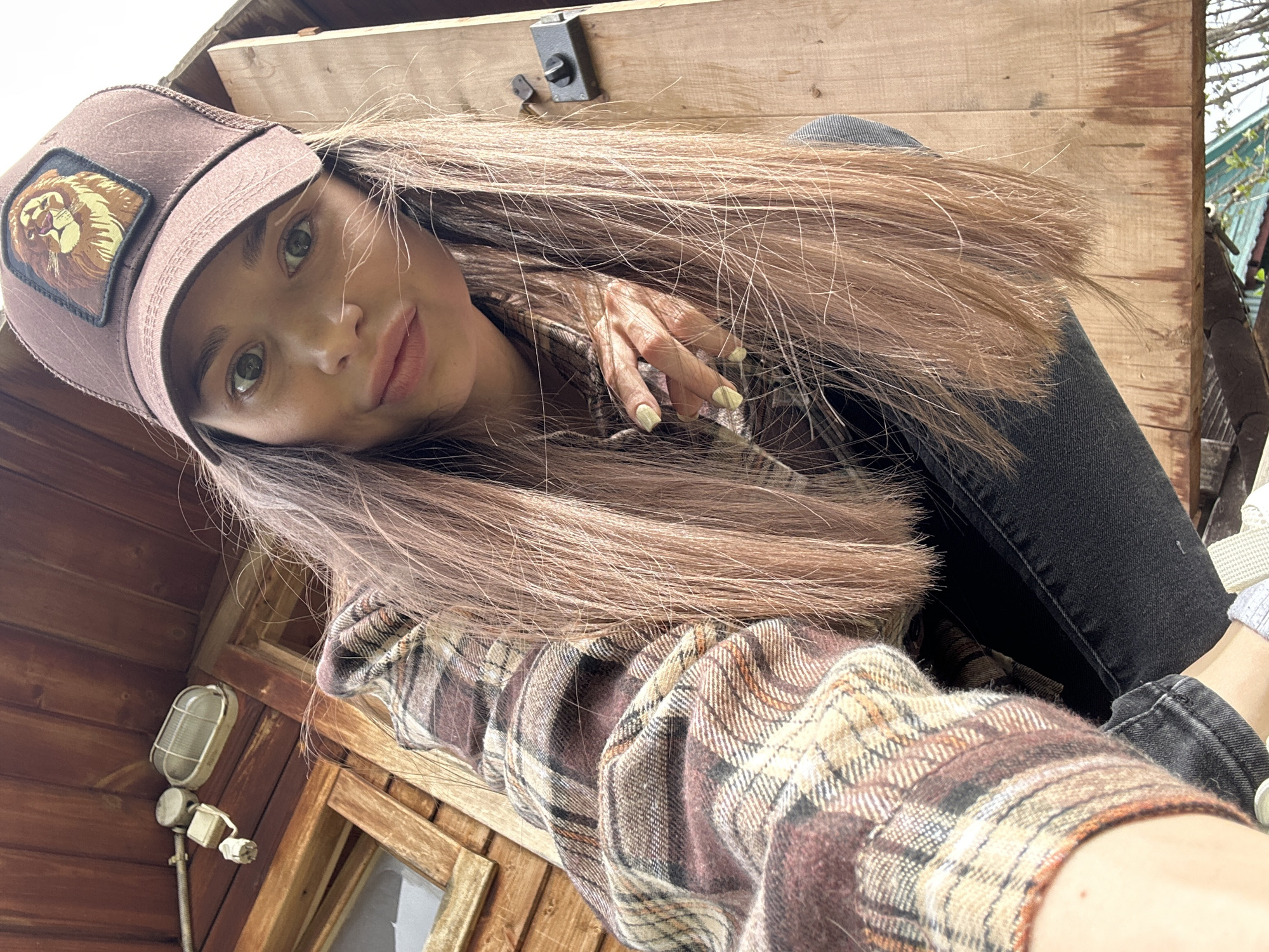 A young woman is posing for a selfie in what appears to be a rustic, wooden cabin or structure, possibly in Khokhryaki, Russia. She is wearing a brown baseball cap with a lion emblem, a plaid flannel shirt, and dark jeans. The lighting is natural, and the background includes wooden walls, a window, and what seems to be part of the cabin’s roof. The woman's hair is long and brown, and her nails are painted a light color.
