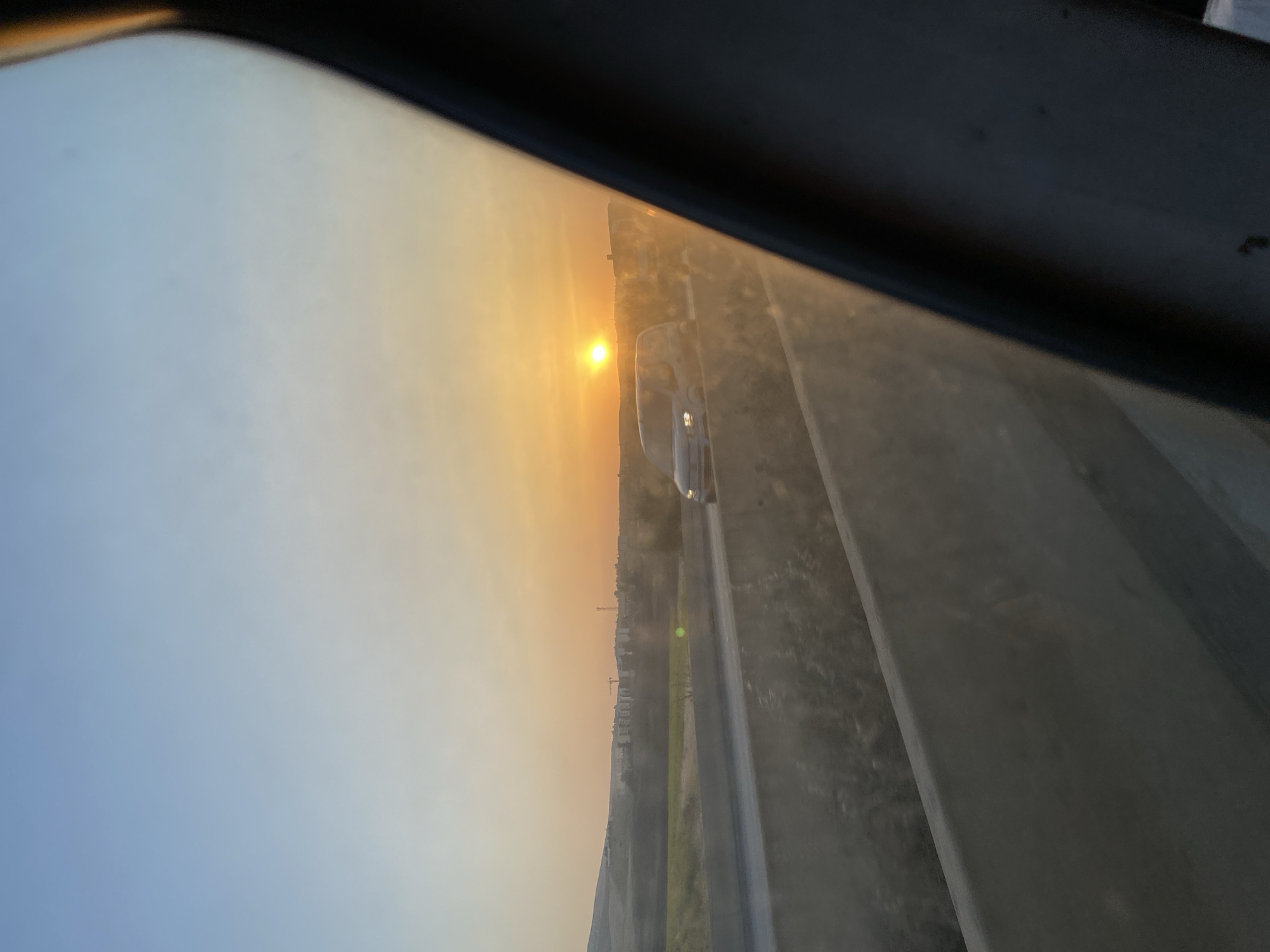 This is a photograph taken from inside a vehicle, looking out at a highway at sunset. The sky is a gradient of blue to orange, with the sun low on the horizon. There are cars on the highway, and in the distance, there are buildings and hills. The scene is likely in or near Ouled Djilali, Algeria, given the context. The foreground is dominated by the interior of the vehicle, with a dark, curved edge on the right side of the frame, likely a car window frame. The middle ground shows a concrete divider on the highway, and beyond that, the road with vehicles. The background features a rural landscape with some development. The overall impression is one of travel at dusk, with the warm glow of the setting sun illuminating the scene.