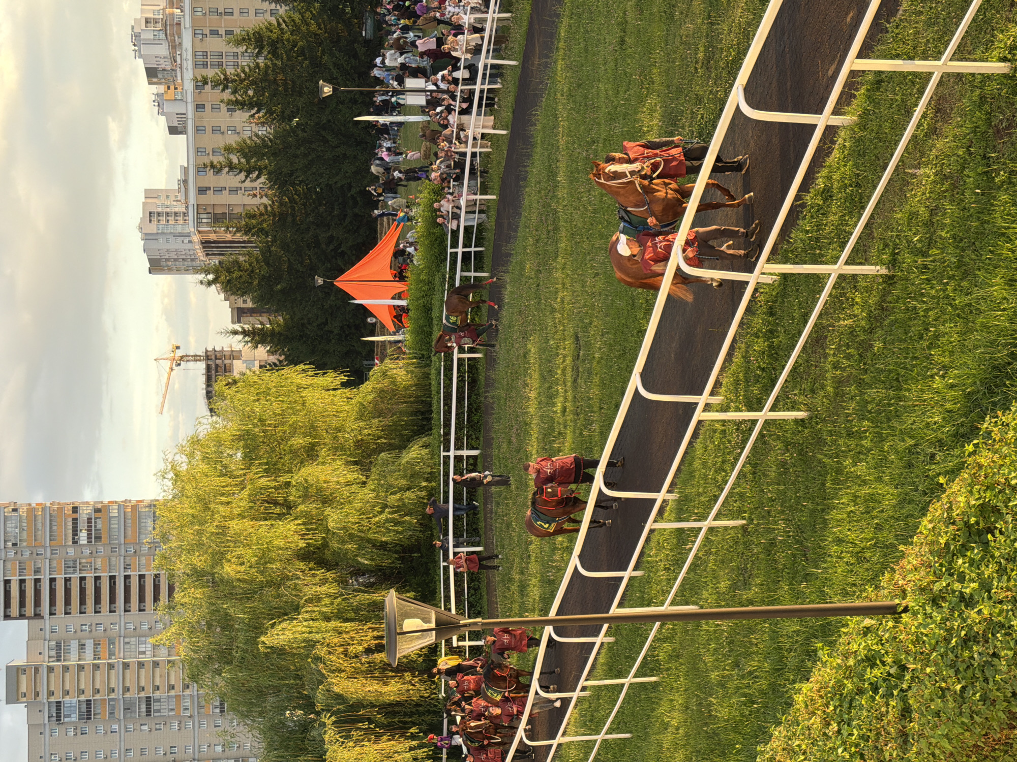 In a sun-drenched outdoor scene, a group of horses and their handlers are parading along a dark asphalt track bordered by a white railing. The handlers are dressed in matching red vests and dark pants, guiding the horses which are adorned with decorative blankets. Behind the track, a lush green grassy area is visible, interspersed with trees and bushes. In the background, a crowd of spectators lines the perimeter, some standing behind another white railing. A large, bright orange tent is set up further back, amidst a cluster of evergreen trees. Dominating the skyline are several multi-story residential buildings, with a construction crane visible in the distance, suggesting an urban or suburban setting. The sky above is partially cloudy with a warm, golden light, indicative of either sunrise or sunset.