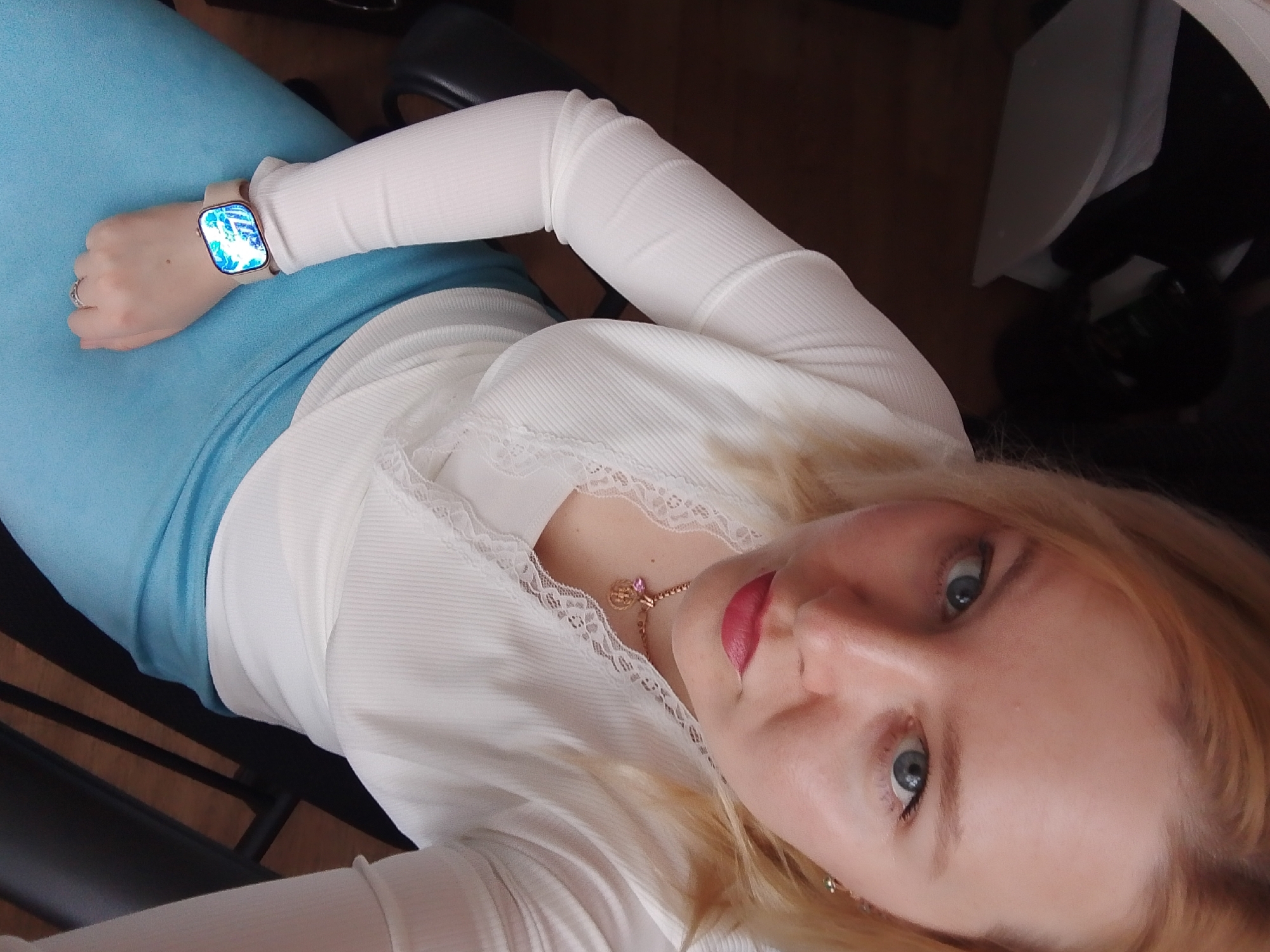 A person is seated on a black chair, likely indoors. They are wearing a white, long-sleeved top with lace trim and a light blue skirt. The person has blonde hair, blue eyes, and is wearing pink lipstick. A smartwatch is on their left wrist. No information indicates a connection to Izhevsk, Russia.