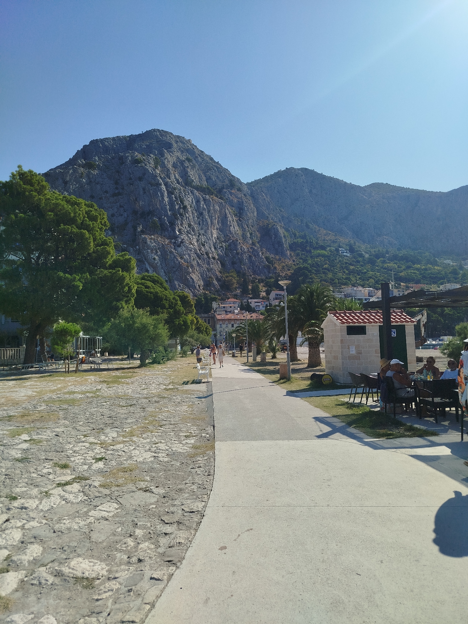 A scenic view of a paved walkway alongside a rocky, tree-lined area, leading towards a town nestled at the base of a large, imposing mountain. The walkway is bordered by a grassy area on one side and a more textured, stone-paved area on the other. Several individuals are seen strolling along the walkway and seated at tables outside a small building with a tiled roof, likely a cafe or restroom facility. Palm trees are scattered throughout the scene, adding to the Mediterranean atmosphere. Buildings with red-tiled roofs are visible in the background, indicating a town or village. The mountain dominates the skyline, its rugged terrain and vegetation suggesting a natural, mountainous landscape. The sky is clear and blue, with the sun casting shadows. The overall scene depicts a leisurely day in a coastal town with a prominent natural backdrop. This is likely the promenade in Omiš, Croatia, known for its dramatic mountain scenery and proximity to the Cetina River.
