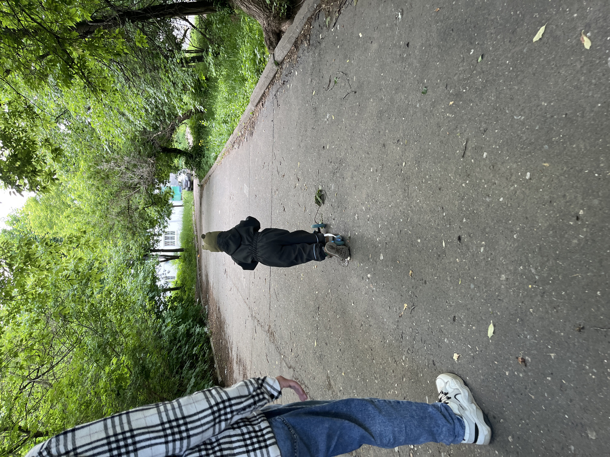 The image captures an outdoor scene from a slightly elevated, first-person perspective, looking down a paved path. In the foreground, the lower half of a person is visible, wearing blue jeans, a black and white plaid shirt, and white sneakers with a dark swoosh-like logo. Further down the path, a child dressed in a dark (possibly black or dark green) full-body suit and a matching hat is seen pushing a blue scooter.
The path is made of asphalt and shows some cracks and debris like fallen leaves and twigs. It is bordered by a curb on the left side, beyond which lies abundant green foliage, including tall trees and dense bushes, suggesting a park or a tree-lined avenue. More greenery is visible on the right side of the path as well. In the distant background, partially obscured by trees, light-colored buildings can be seen, hinting at a residential or urban area.
The lighting suggests it is daytime, possibly under an overcast or partly cloudy sky, as there are no harsh shadows. The overall impression is one of a leisurely outdoor activity. The location context provided is Chaikovsky, Russia. There is no discernible text other than a potential brand logo on the sneaker.