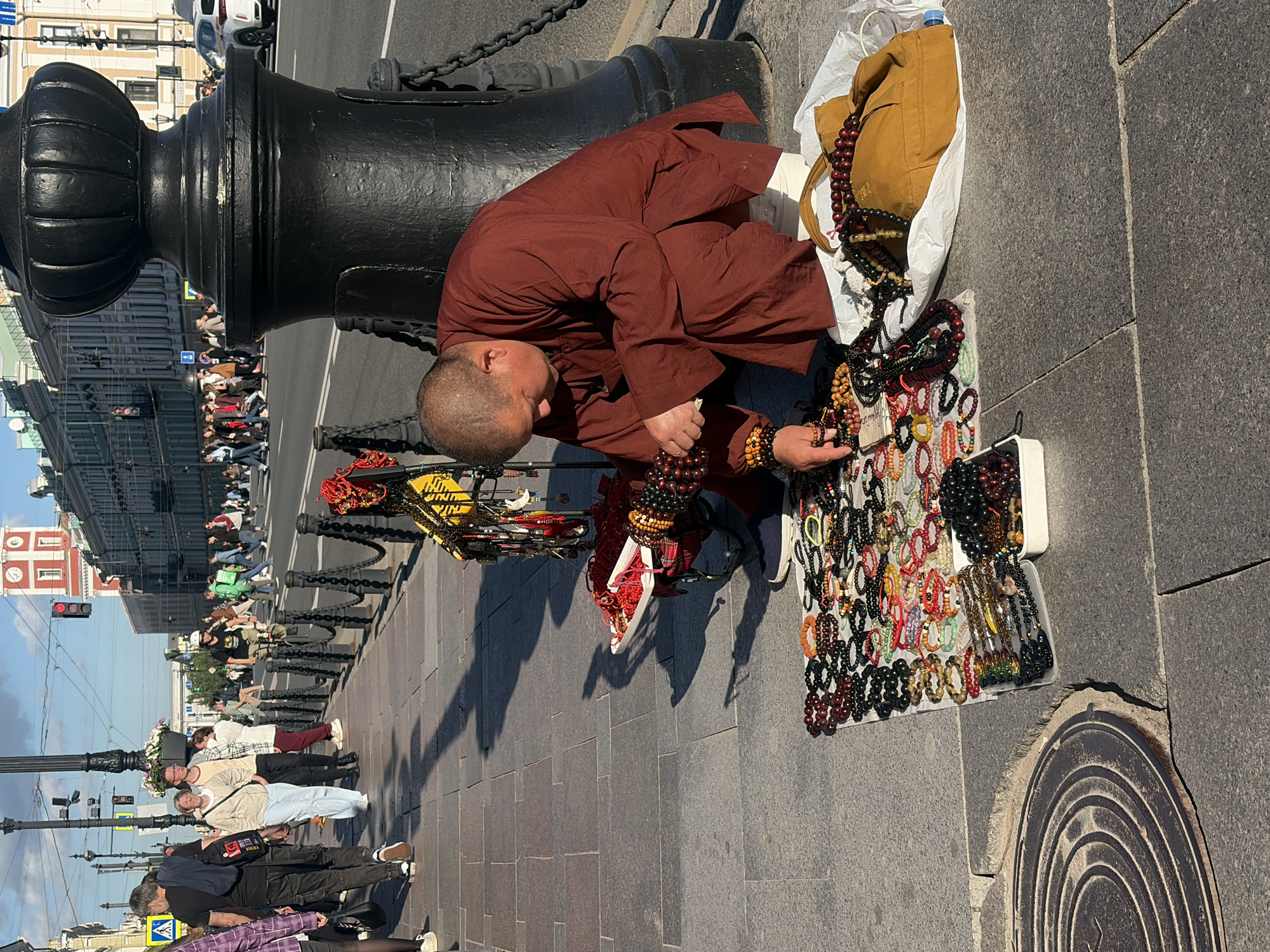 The image shows a street scene in Saint Petersburg, Russia, likely on Nevsky Prospekt, a major thoroughfare known for its historic architecture and bustling atmosphere. In the foreground, a man dressed in a maroon robe is crouched on the sidewalk, arranging a display of prayer beads and bracelets. The items are spread out on a white cloth and in white trays. He is actively engaged in his work, with his hands manipulating the beads.
Behind him, a large, ornate black lamppost stands prominently, casting shadows on the stone pavement. The sidewalk is wide and paved with large, grey stone slabs, with a circular metal grate visible in the lower left corner. In the mid-ground, pedestrians are walking along the sidewalk and crossing the street. Some are casually strolling, while others appear to be shoppers. The background features a row of grand, historic buildings, characteristic of Saint Petersburg's urban landscape. A red-and-white clock tower is visible in the distance. The sky is blue with some clouds, suggesting a pleasant day. The presence of a road with vehicles and streetlights indicates an urban environment.