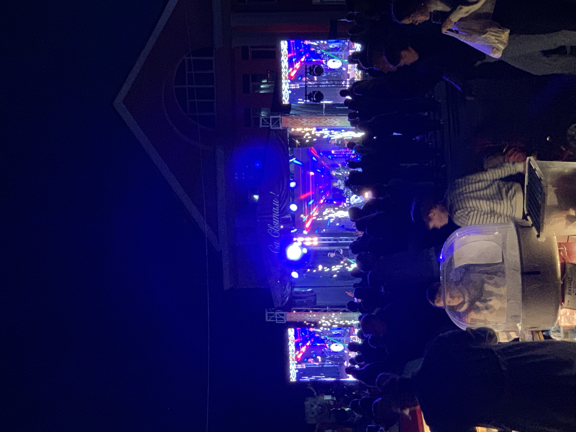 The image depicts an outdoor evening event, likely a concert or festival, in the city of Kapyĺ, Belarus. In the background, a building with a distinctive arched window is illuminated by the stage lights. The stage itself is brightly lit with a band performing and a large banner overhead that appears to have text written on it. The foreground is filled with a crowd of people silhouetted against the stage, observing the performance. To the left, a person is operating a cotton candy machine, with a child visible inside its transparent enclosure. Several other individuals are standing around, some holding bags. The overall atmosphere suggests a lively public gathering.