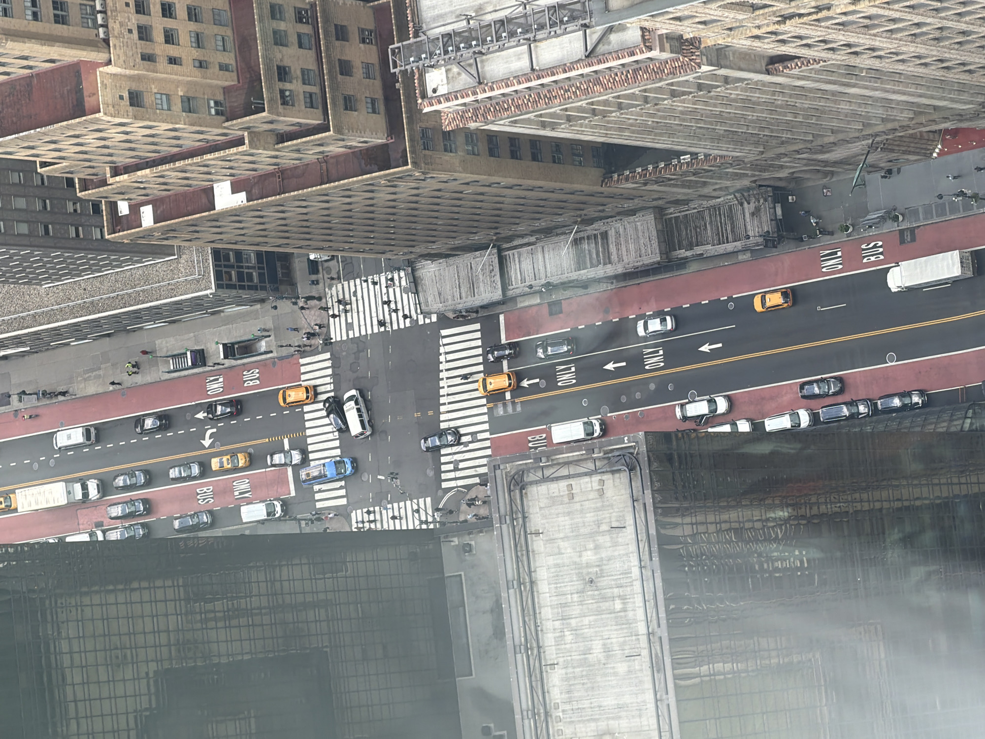 This is an aerial view of a street in New York City, United States of America. The street has a dedicated bus lane marked in red with "BUS ONLY" painted on the asphalt. Several vehicles are visible, including cars, a bus, and a truck. Pedestrians are seen walking on the sidewalks and crossing the street at a crosswalk. The buildings lining the street are typical of New York City, with some appearing to be older brick structures and others more modern glass-fronted buildings. The sky appears overcast, and the lighting suggests it is daytime.