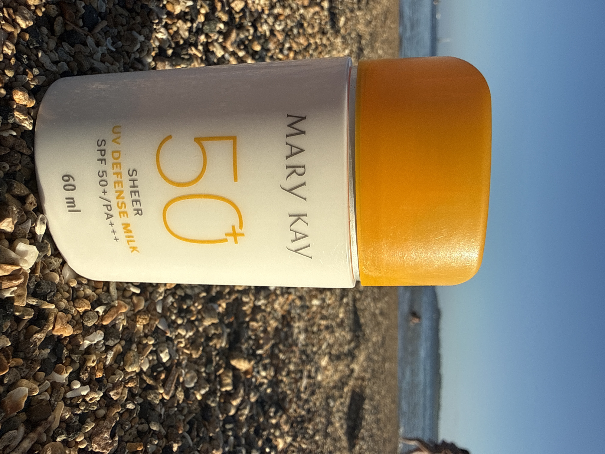 A bottle of Mary Kay SPF 50+ UV Defense Milk sits on a pebbled beach. The bottle is white with an orange cap and features orange text with "50+" prominently displayed. In the background, the ocean meets a clear blue sky, with a few blurry figures visible near the water. The location is the city of Denpasar, Indonesia.