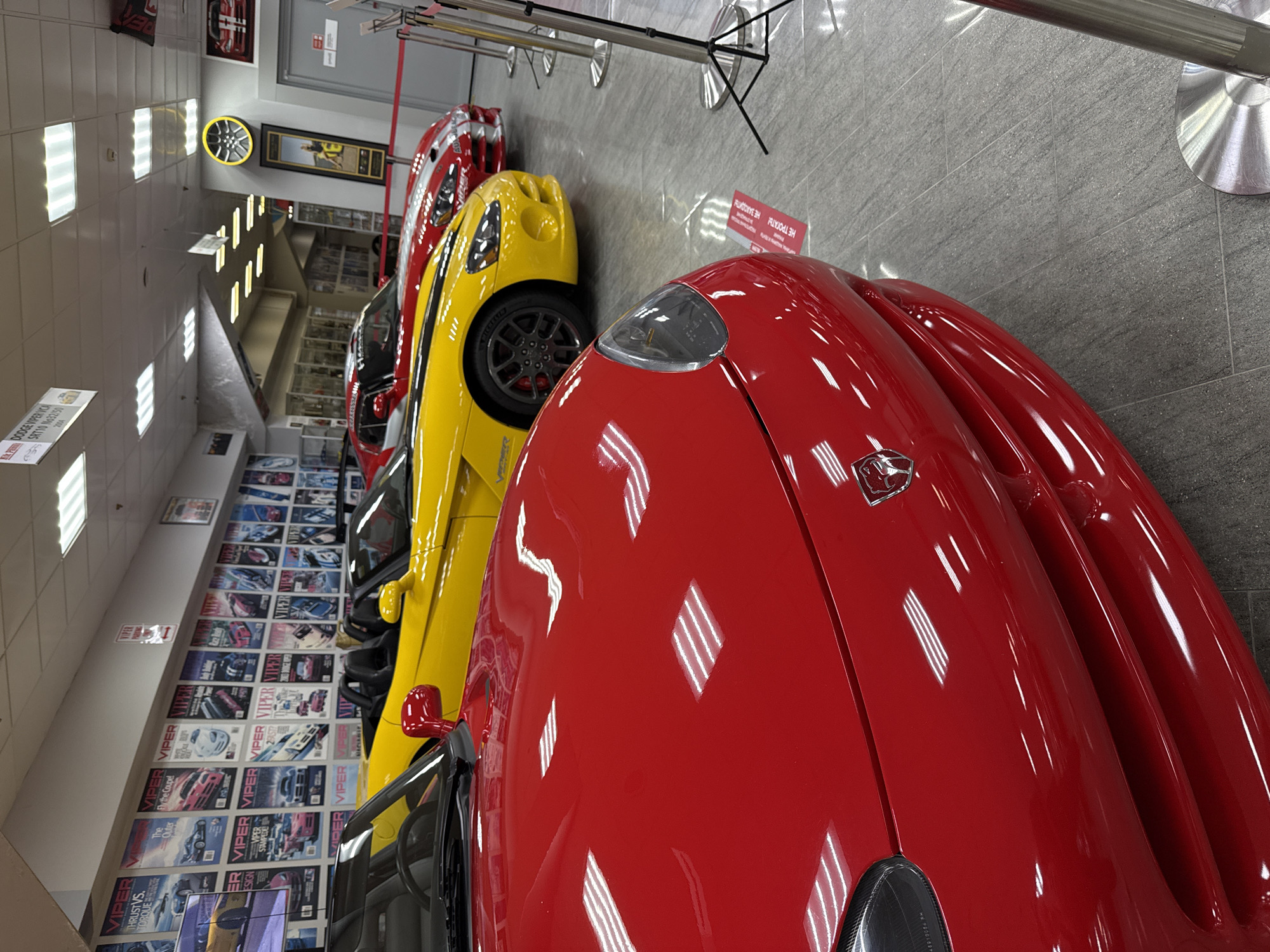 The image depicts an indoor display of several high-performance Dodge Viper sports cars, meticulously lined up in what appears to be a museum or showroom, located in Sirius, Russia. In the foreground, a glossy red Dodge Viper, possibly a coupe, is prominently featured, with its sleek hood, headlight, and a partial view of its front fender. The car's surface reflects the bright, rectangular fluorescent lights on the ceiling. A "VIPER" logo is visible near the front fender.
Immediately behind the red car is a vibrant yellow Dodge Viper, showing its front-left wheel with "MICHELIN" branding on the tire, and its side profile. Further down the line, partially obscured, is a red and white striped Viper.
The setting features light grey, possibly tiled, walls and a reflective grey floor. The walls are extensively decorated with framed posters or magazine covers, all centered around the Dodge Viper, showcasing various models and headlines such as "THRUST VS. TORQUE," "The Outer," "Fly the Coupe," and "VIPER DESIGN." One poster specifically mentions "DODGE VIPER SRT10 No32/50."
A metal barrier with stanchions separates the viewer from the cars, indicating a restricted access display. A red sign attached to this barrier clearly states in Russian: "НЕ ТРОГАТЬ НЕ ЗАХОДИТЬ," which translates to "DO NOT TOUCH, DO NOT ENTER" or "NO ENTRY." Other wall decorations include a black-framed picture and a circular object, possibly a wheel rim, illuminated by a yellow halo. The lighting is bright and even, emanating from numerous rectangular overhead fixtures. No people are visible in the scene.