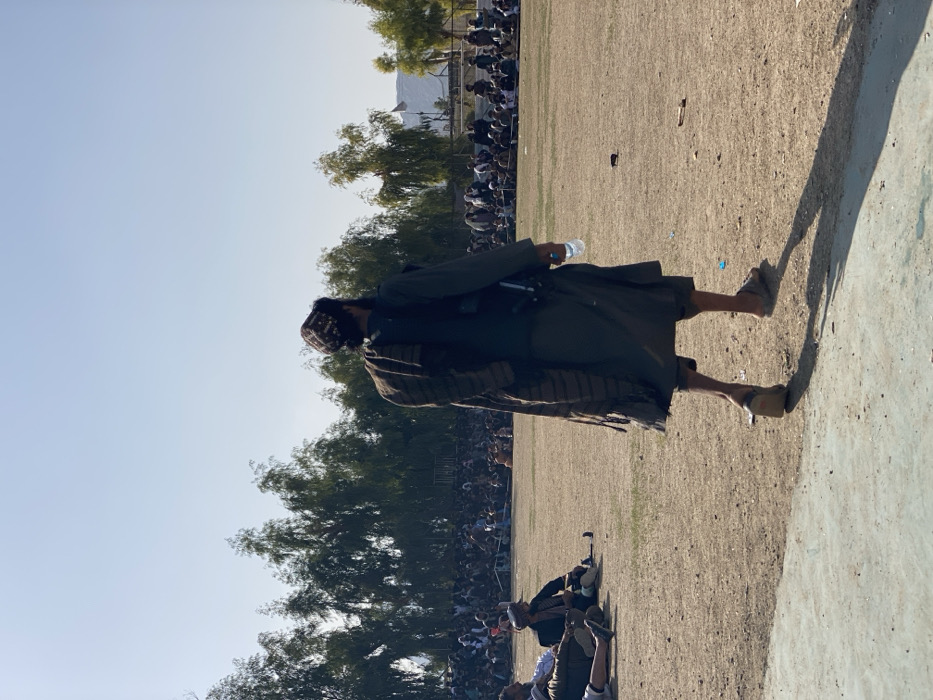 An outdoor scene in Kandahar, Afghanistan, under a clear sky, depicting a large gathering. A male figure, viewed from the back, walks rightward across a dry, unpaved dirt field. He wears a dark tunic, dark vest, patterned skullcap, and sandals, with a striped scarf draped over his left shoulder. He holds a clear plastic water bottle in his right hand. A dark, long-barreled weapon is slung across his back, partially visible beneath the scarf and vest. His long shadow projects behind him to the left. In the midground, to the left, several men are seated on the ground, with one individual lying down. In the background, a large assembly of men is seated directly on the ground, forming an arc-like arrangement, observing the field. Behind the crowd, a dense line of tall, leafy trees provides a green backdrop. A white flag on a pole is discernible in the distant right background, beyond the trees.