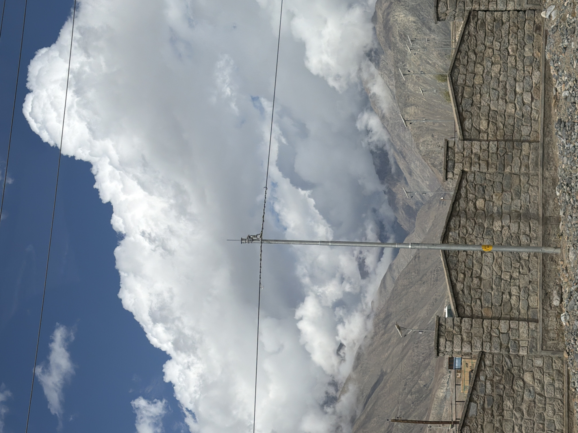 The image captures a mountainous landscape near Spituk, India, under a partly cloudy sky. A dry, rugged mountain range dominates the background, with barren slopes and shadowed peaks. In the foreground, a sturdy stone wall constructed from irregular, weathered stones forms a barrier, with a textured concrete base. Several tall, thin metal poles with what appear to be solar panels or lighting fixtures are strategically placed along the top of the wall, suggesting infrastructure development. A prominent silver pole with some electrical equipment stands near the center of the frame. Overhead, power lines stretch across the scene, silhouetted against the bright sky and dramatic white clouds. The ground in front of the wall is covered with gravel and smaller stones. The overall impression is one of a remote, possibly developing area, utilizing solar technology amidst a harsh, natural environment.