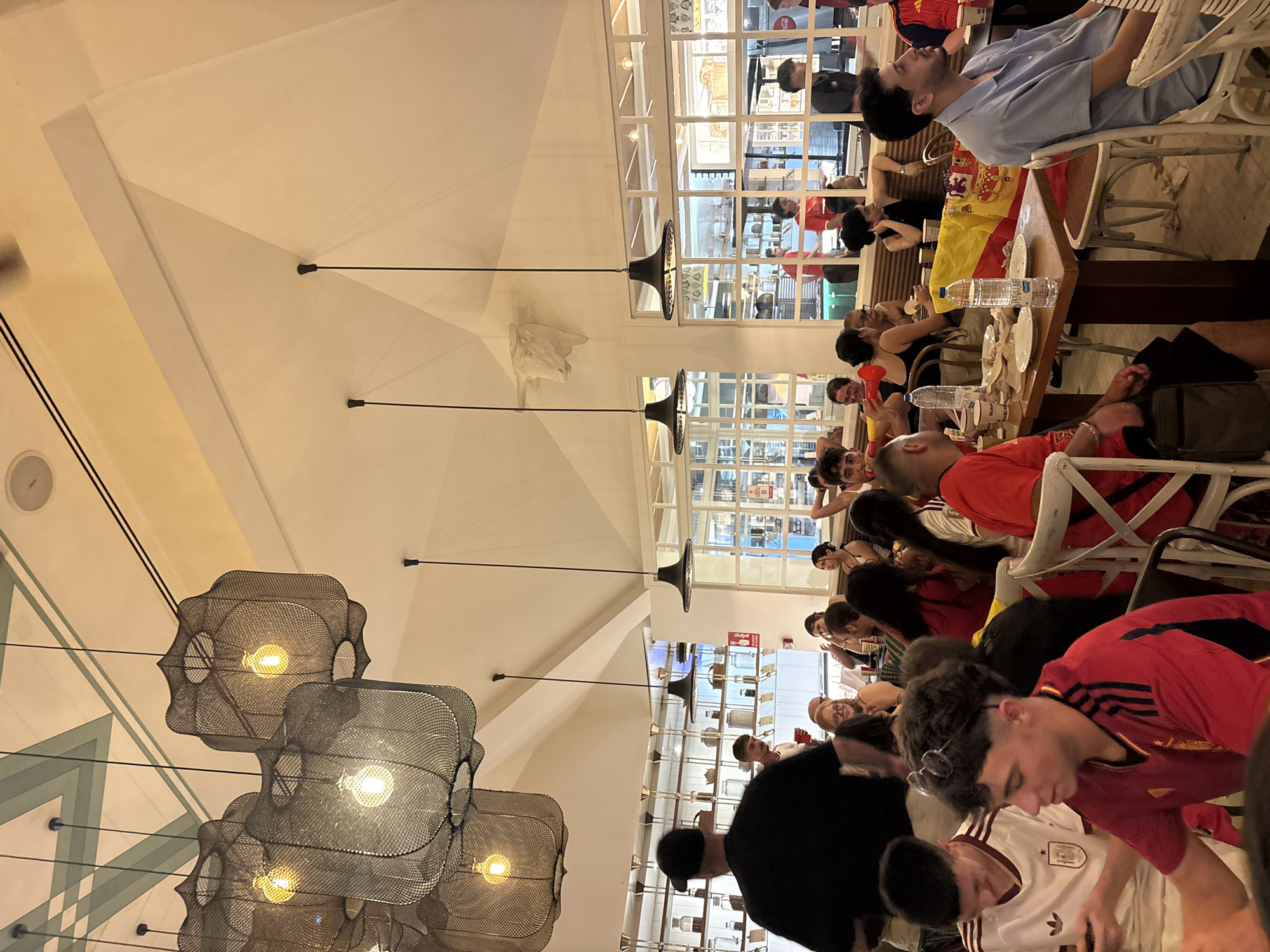The image depicts the interior of a restaurant in València, Spain, likely a casual dining establishment or tapas bar, filled with patrons. The architecture features high ceilings with exposed beams and a distinctive geometric ceiling design in shades of white and pale green. Several large, intricately woven pendant lights hang from the ceiling, casting a warm glow. In the background, a decorative bull's head sculpture is mounted high on the wall.
The restaurant is busy with people seated at tables, engaged in conversation and dining. Many patrons are wearing Spanish national football team jerseys, suggesting a sporting event is being celebrated or watched. The tables are set with plates, cutlery, bottles of water, and food. A Spanish flag is prominently displayed on one of the tables.
In the foreground, several young men are visible. One, wearing a red Spanish jersey, is looking to his right, holding a horn. Another, in a white jersey, is looking down at his phone. Behind them, a man in a blue shirt is seated at a table. Various other individuals and groups are scattered throughout the dining area, their faces obscured or partially visible. The overall atmosphere appears lively and social. Shelving units displaying bottles and pottery are visible on the walls in the middle ground. Large windows, framed in white, allow light to enter and provide glimpses of what appears to be a busy street or another part of the establishment.