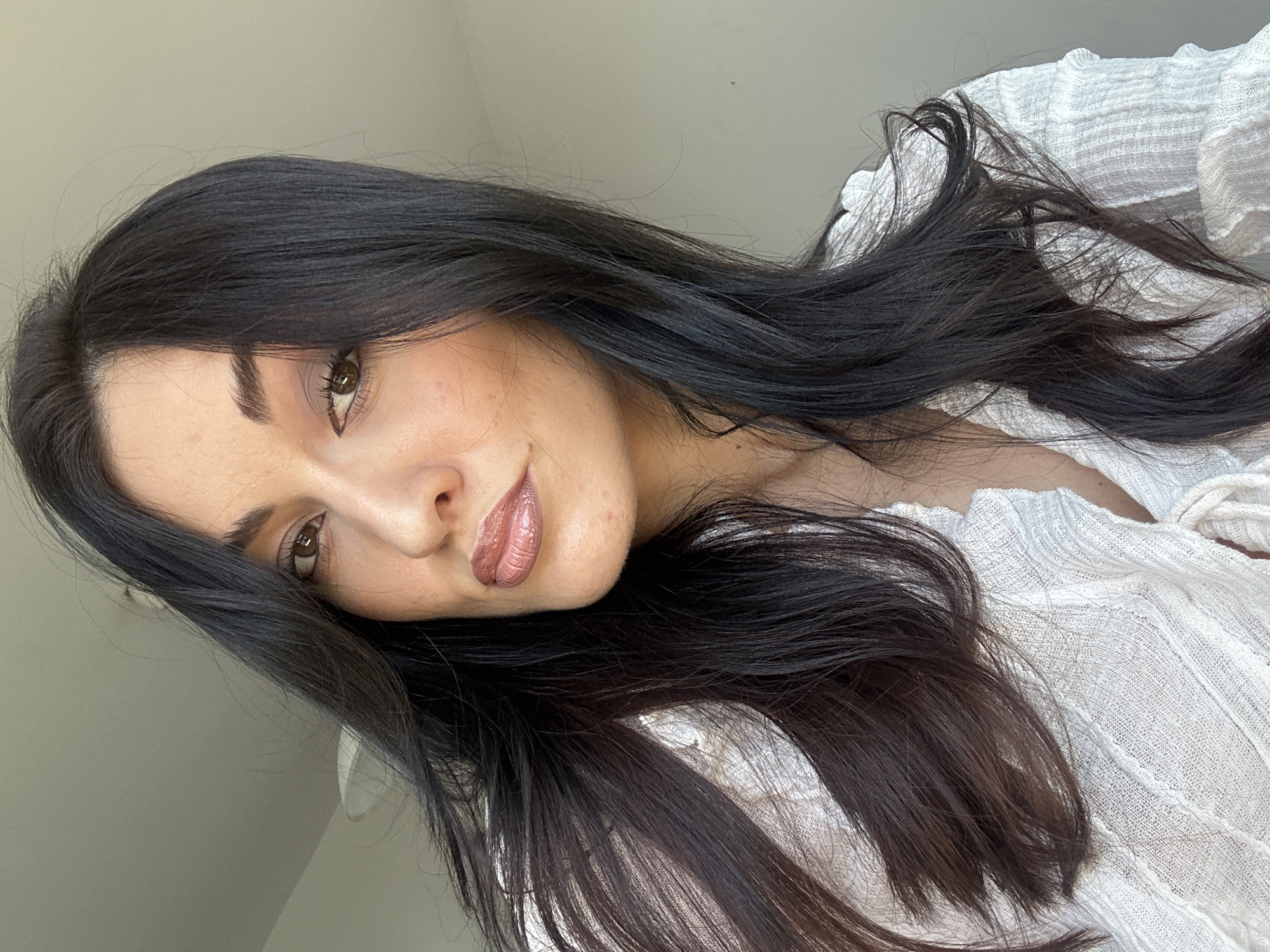 The image shows a close-up, head-and-shoulders portrait of a woman with long, dark hair. She is wearing a white, textured top with ruffled details around the neckline. Her makeup includes subtle eye shadow and a shimmery, nude-toned lipstick. The background is out of focus, appearing to be an indoor setting with a light-colored wall and ceiling. There is no visible indication of the city of Burgas, Bulgaria, or any specific urban environment.