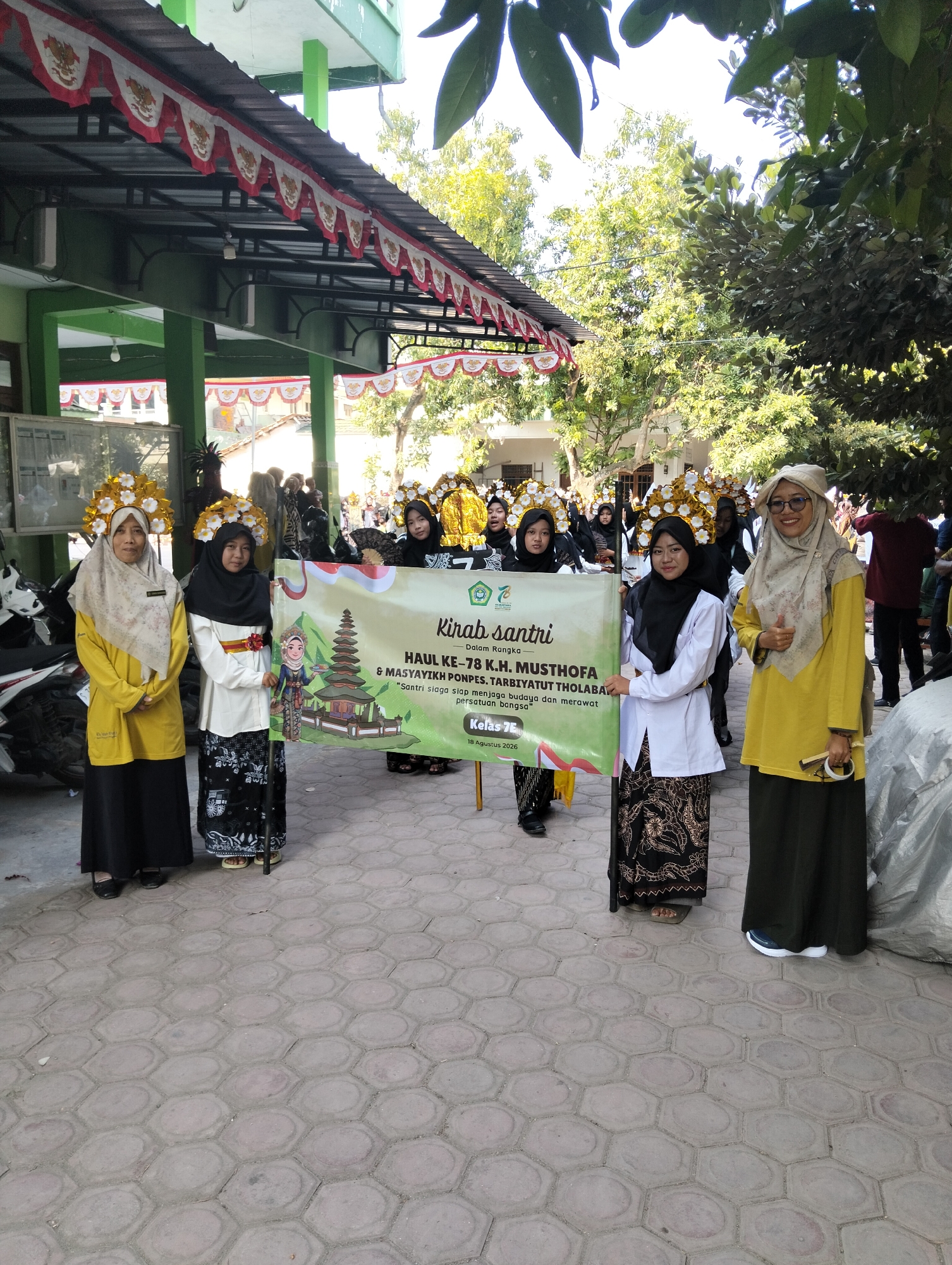 In Kranji, Indonesia, a group of people, predominantly young women and a few adults, are participating in an outdoor event. The individuals are dressed in traditional Islamic attire, with many of the younger participants wearing hijabs and long dresses, some adorned with elaborate headdresses. One young woman in the foreground holds a banner with the text "Kirab Santri" and "HAUL KE - 78 K.H. MUSTHOFA & MASYAYIKH PONPES TARBIYATUT THOLABA". The banner also features an illustration of a traditional Indonesian building and a cartoon character holding a puppet.

Two women in the foreground, one wearing a yellow top and the other a white top with a black hijab, are standing next to the banner. The woman in yellow gives a thumbs-up gesture. Another woman on the far left wears a yellow top and a black skirt. Behind them, a line of participants are visible, some holding flags or banners. The background shows a building with a green facade and a roof decorated with red and white banners. Trees and foliage are visible in the background, suggesting a sunny day. The ground is paved with hexagonal tiles. Several motorcycles are parked on the left side of the image. The overall scene depicts a cultural or religious procession.