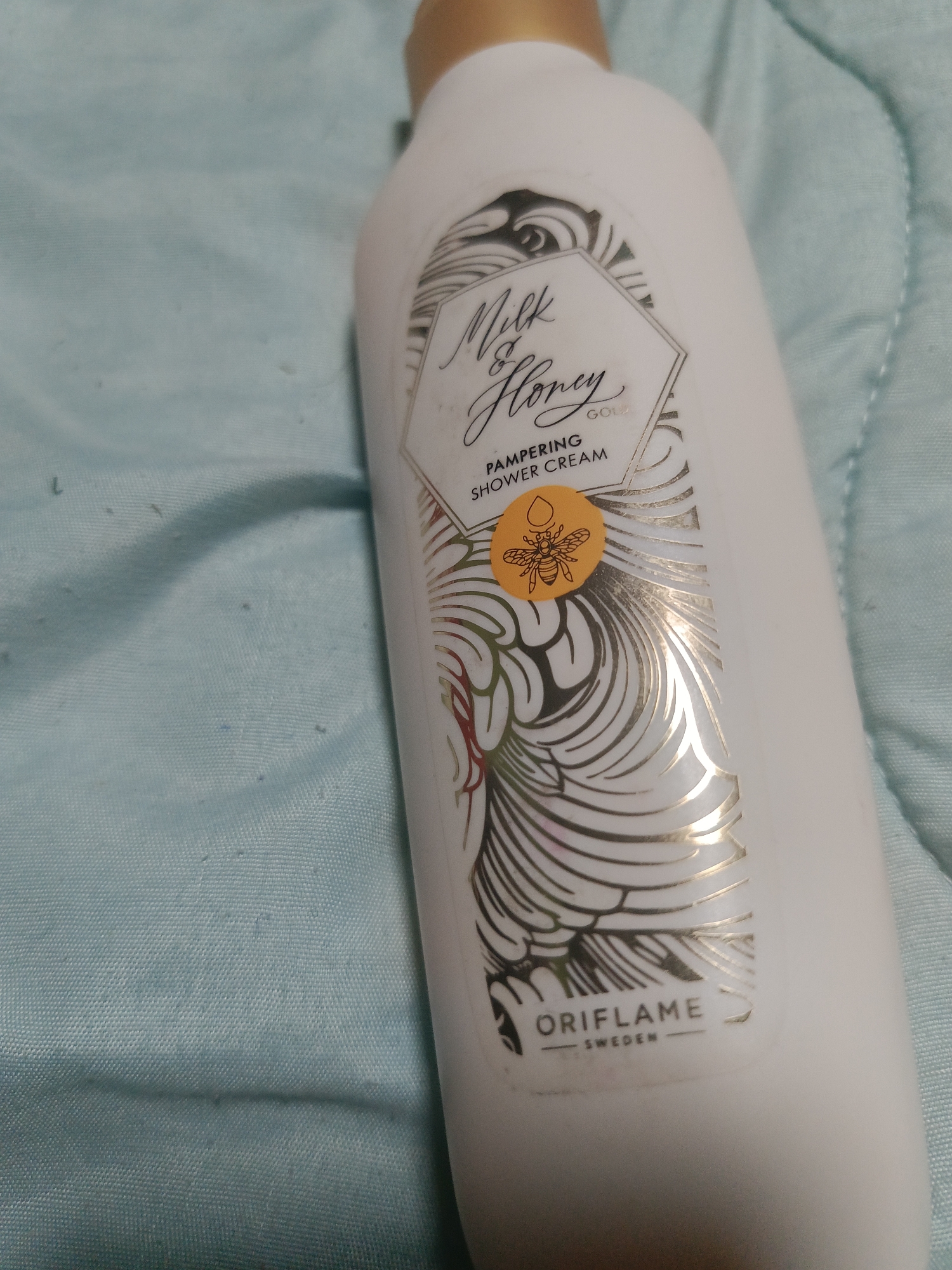 A close-up, slightly tilted, eye-level shot shows a white bottle of Oriflame Milk & Honey Gold pampering shower cream, lying on a textured, light blue fabric surface. The bottle's label features an ornate design with gold accents, a hexagonal frame, and text that reads "Milk & Honey GOLD," "PAMPERING SHOWER CREAM," and "ORIFLAME SWEDEN." A small, circular sticker with a bee illustration is prominently displayed on the label. The fabric surface is slightly wrinkled and shows small, dark speckles. The background is blurred, with soft lighting. There are no people present in the image.