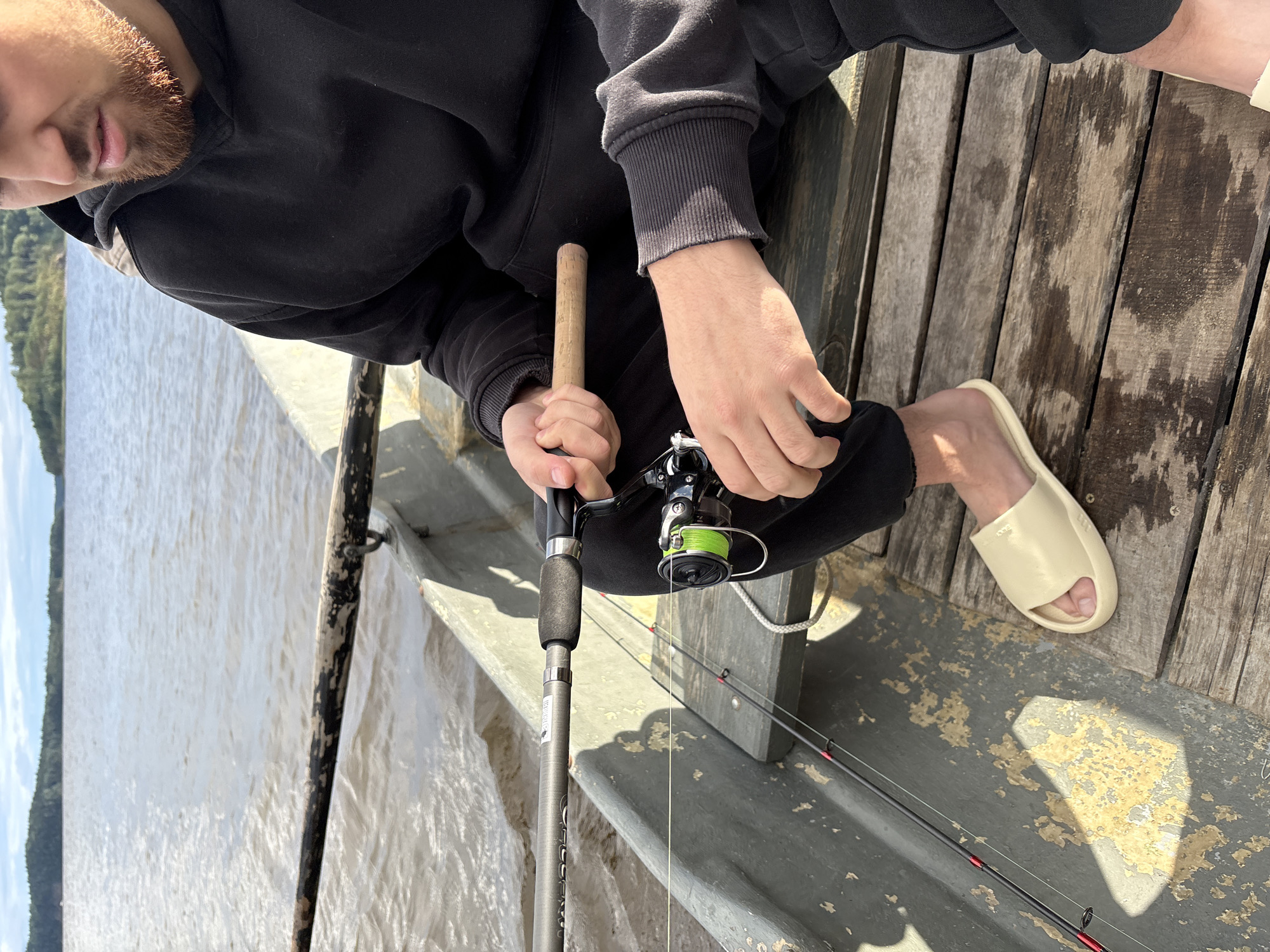 A person wearing a black hoodie and black pants is sitting in a boat on a body of water. They are holding a fishing rod with a green reel. The boat is made of metal and has wooden seating. In the background, there is a shoreline with trees and a cloudy sky. The location is identified as Rautalakhti, Russia.