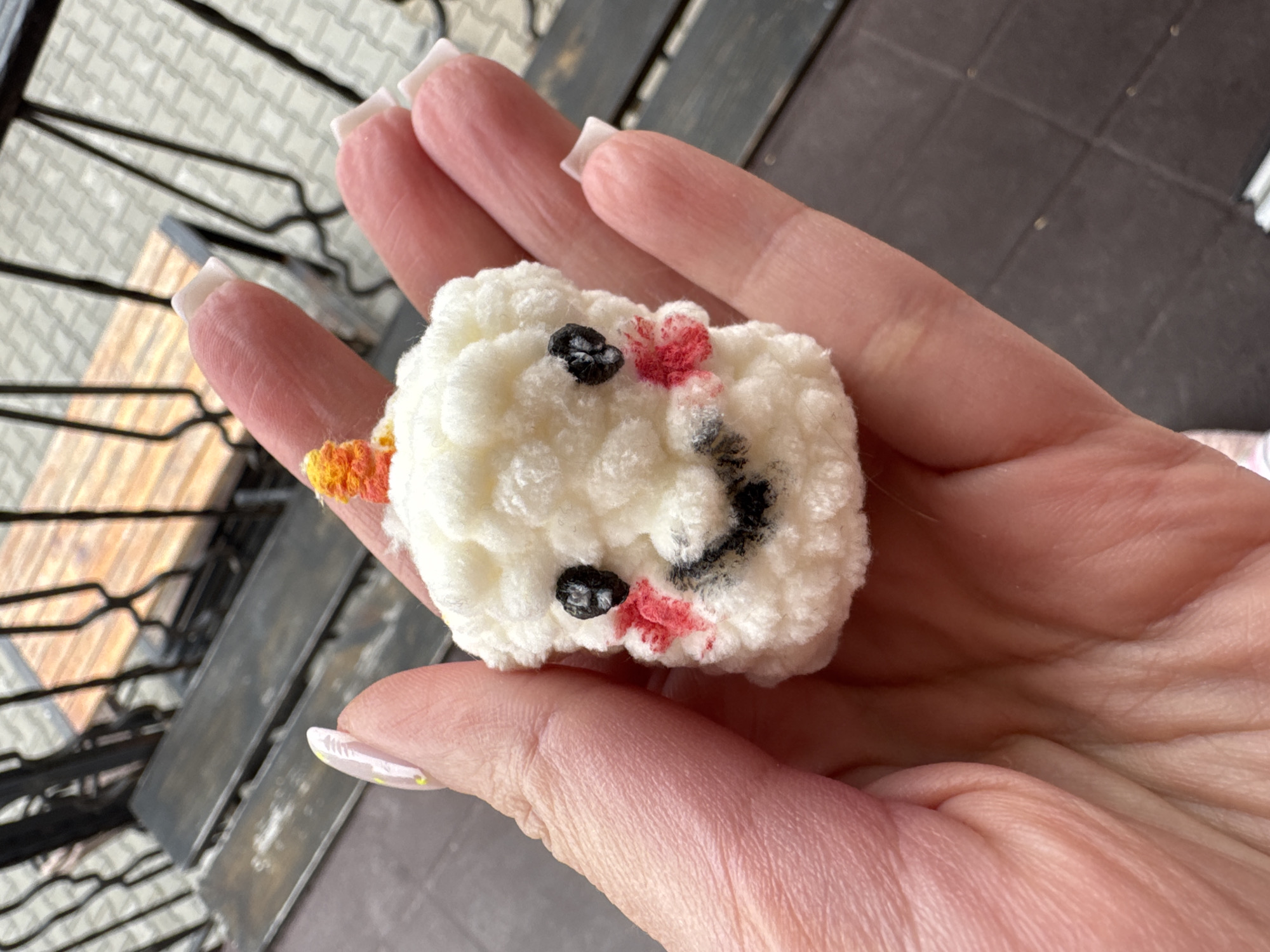 A hand holds a white, square-shaped, crocheted object resembling a face with black eyes, a black smiling mouth, and red cheeks. The person has light skin with a short, square white manicure. A balcony or deck with a metal railing is in the background. Based on the metadata, the location is Ждановіцкі сельскі Савет, Belarus.