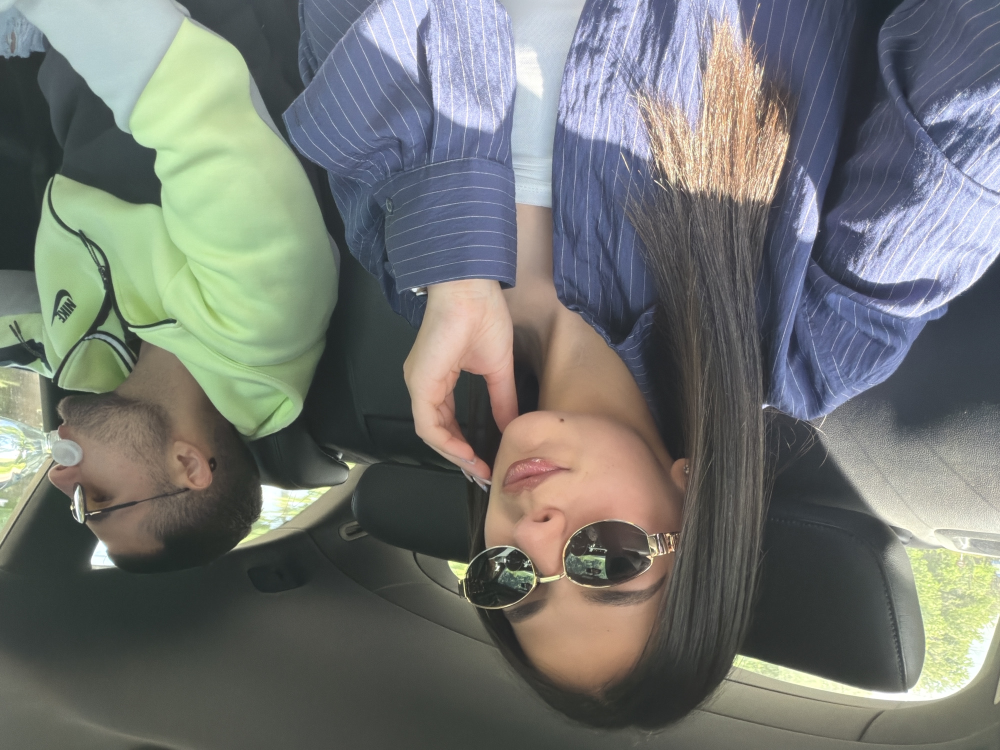 The image shows two individuals inside a car. A woman with long dark hair and gold-rimmed sunglasses is in the foreground. She is wearing a blue and white striped shirt. A man with glasses, a beard, and wearing a light green Nike sweatshirt is visible in the background. He is holding a water bottle in his mouth. The location cannot be determined from the image.