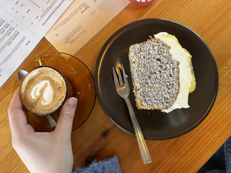 An overhead photograph depicts a tabletop scene inside a cafe or restaurant located in Kaiserslautern, Germany. In the upper left quadrant, a human hand, presumed female, holds an amber-tinted glass cup containing a latte with heart-shaped art on its foam. The cup rests on a matching saucer, which also holds a small silver spoon. A portion of a blue knitted garment is visible on the left, belonging to the individual whose hand is shown.
In the lower right quadrant, a slice of cake is presented on a dark brown, matte ceramic plate. The cake is light greyish-brown with visible yellow flecks, topped with a thick layer of white frosting and a yellow curd or cream layer beneath it. A bite has been removed from the cake's left side. A silver-colored metal fork rests on the plate to the left of the cake.
The table surface is made of light-to-medium brown wood with visible grain. To the right of the food items, a laminated menu in German is visible, showing sections like "WEIN" and "GETRÄNKE & HAUSGEMACHTES" with prices in Euros. Behind this menu, a white sheet of paper confirms the location with "KAISERSLAUTERN" printed at the top, along with other German text including "French Toast" and "CA. 15 UHR". A small, indistinct red object is partially visible on the far right edge of the table.