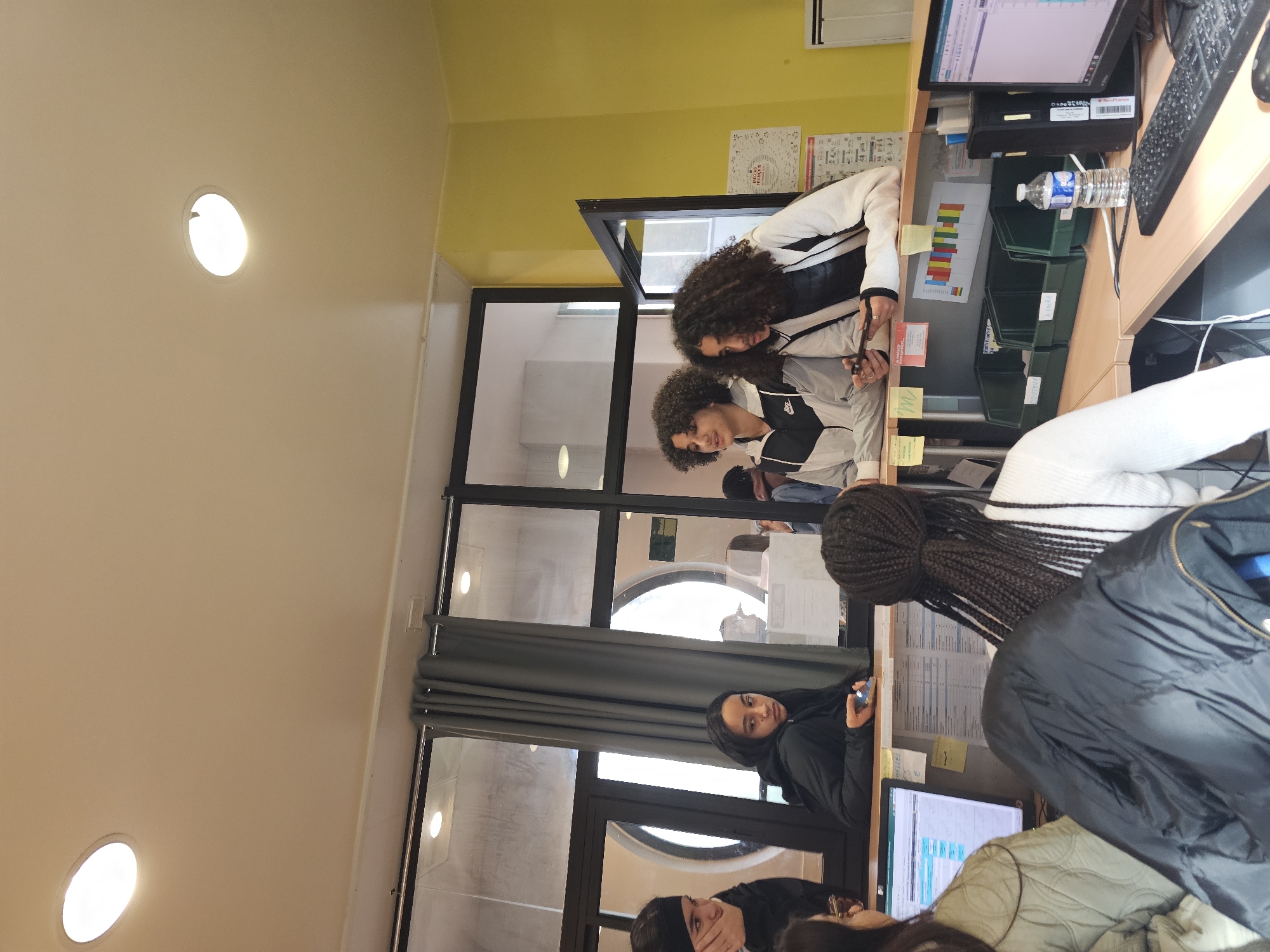 Here is a description of the image:
The image appears to be taken indoors, possibly in an office or communal workspace. A group of individuals are present, some are standing and interacting near a desk with a computer and various office supplies, while others are seated in the foreground. The environment includes large windows providing natural light, with a view possibly showing an urban setting. The color palette is dominated by neutral tones (beige, white, gray) with accent colors (yellow, black). The presence of windows, wall paint, and general aesthetics suggests that the location may be Paris, France.