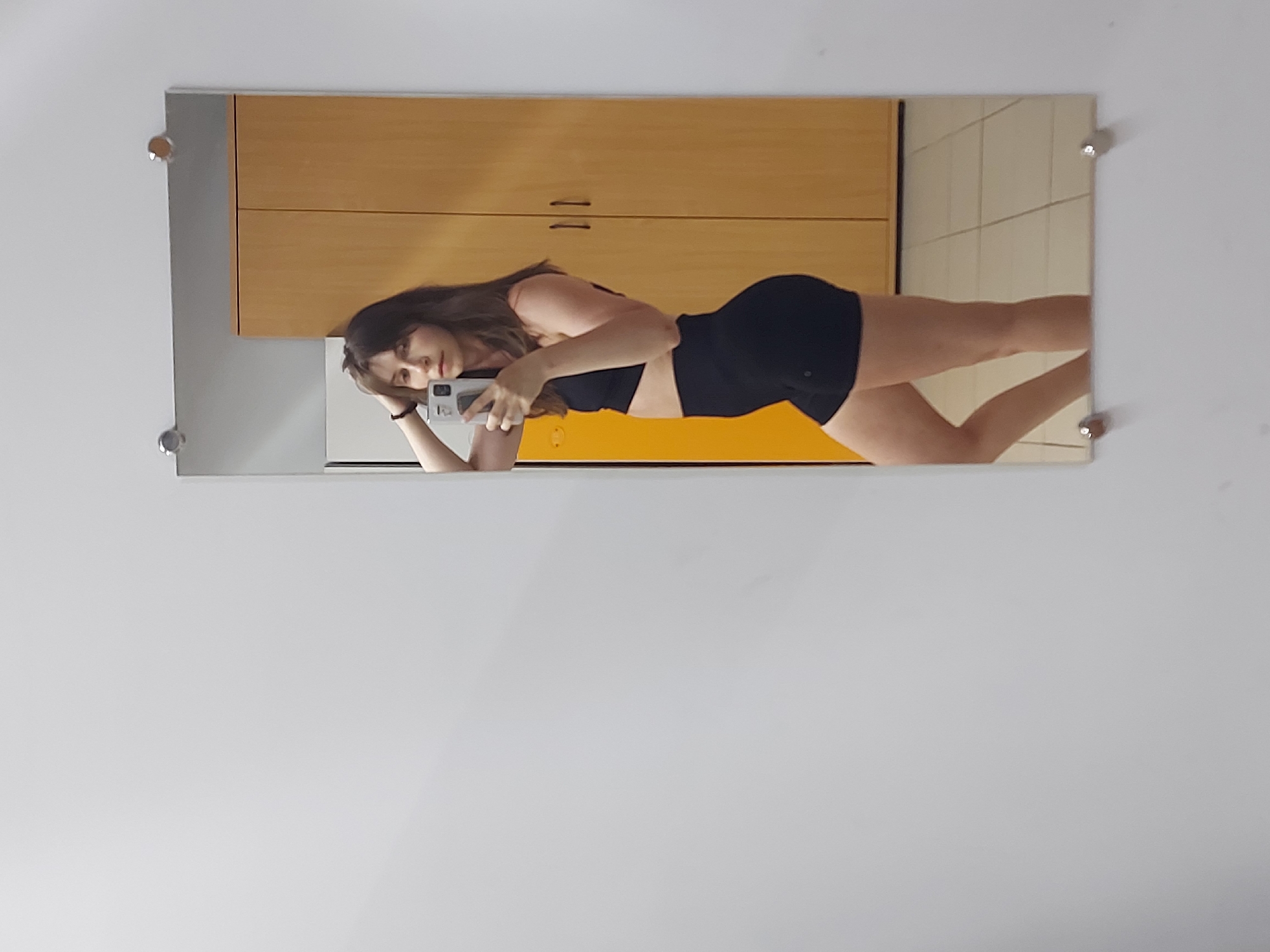 The image shows a reflection in a rectangular mirror mounted on a white wall. The reflection depicts a woman lying down, wearing a black sports bra and black shorts. She is holding a cell phone and appears to be taking a selfie. In the background of the reflection, there are light-brown wooden cabinets and off-white tiles. The location is not specified.
