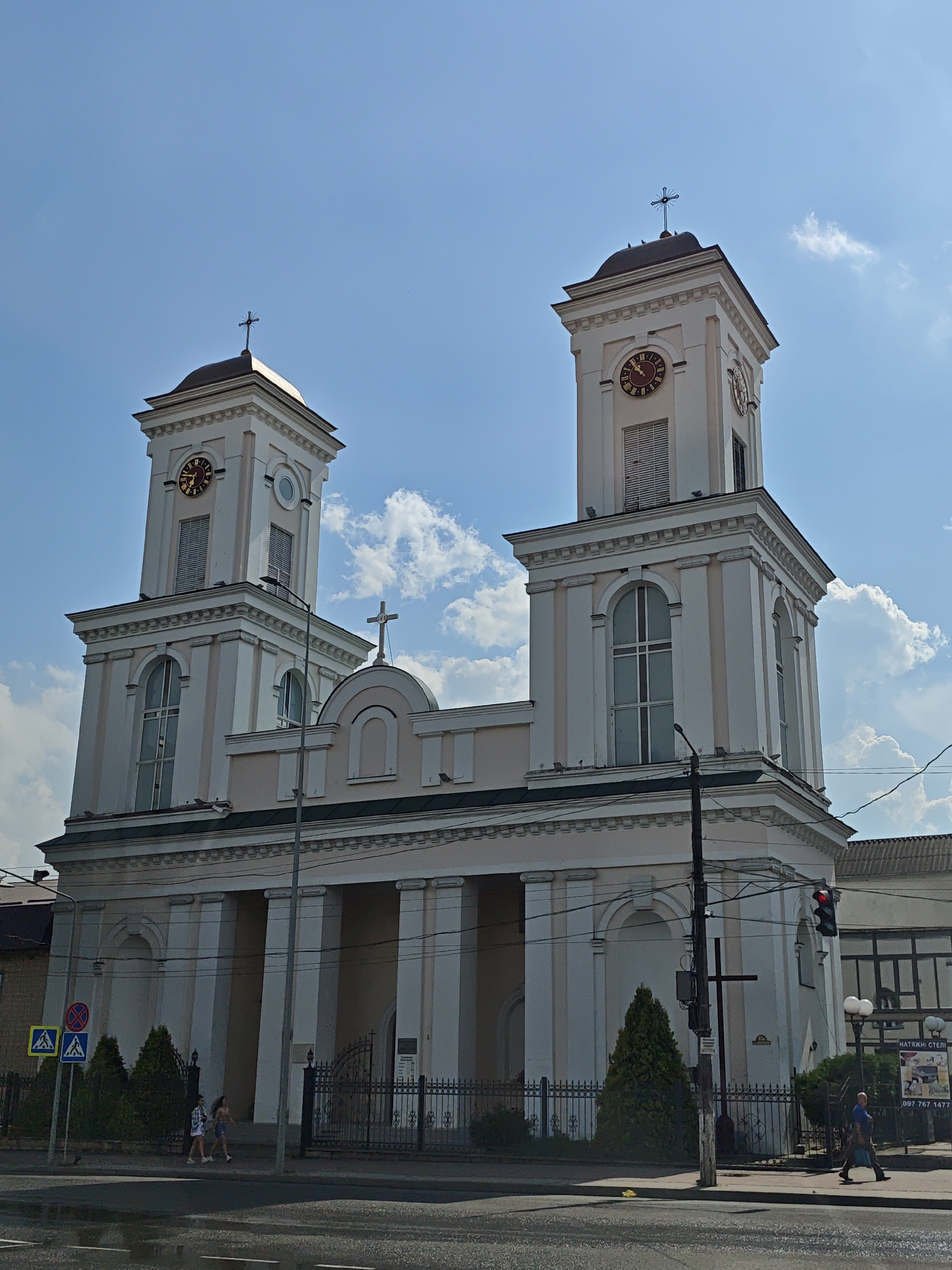 The photograph captures a daytime view of the Church of the Assumption of the Blessed Virgin Mary in Nemyriv, Ukraine. The building features a symmetrical facade with two prominent bell towers, each topped with a cross. Both towers have clocks with Roman numerals, and one clock face is partially obscured. Arched windows and pilasters adorn the towers and the central structure. A portico with Corinthian columns extends across the front of the church, leading to an entrance.
In the foreground, a street with wet asphalt suggests recent rain. Pedestrians walk along the sidewalk in front of the church, with some trees and street signs visible. A traffic light is illuminated red. To the right, a building with a sign in Ukrainian, "НАТЯЖНІ СТЕЛІ", is partially visible. The sky is blue with scattered clouds, and sunlight illuminates the scene.
