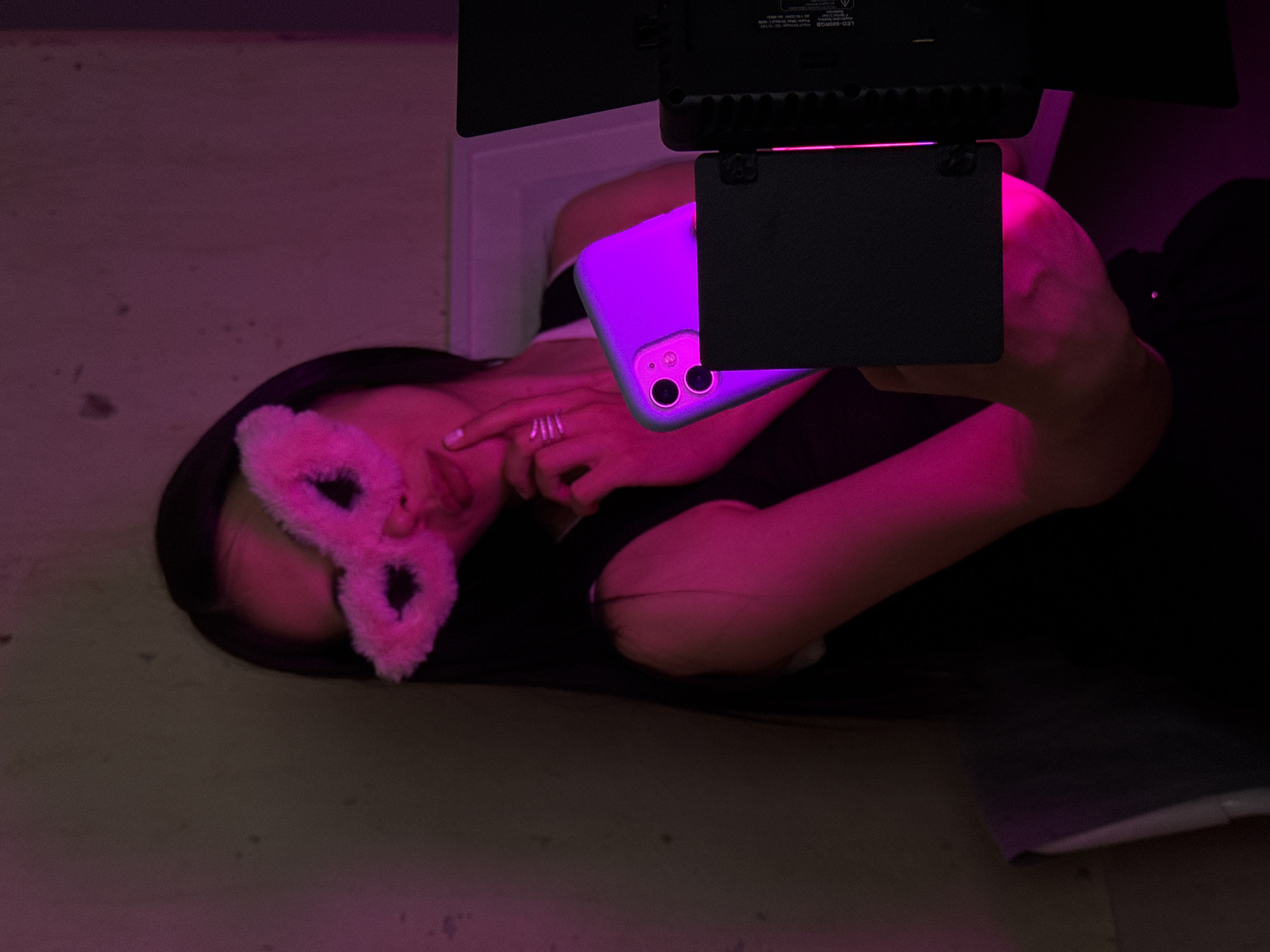 An individual, partially obscured by a fluffy pink mask resembling bunny ears and illuminated by purple light, is seen holding a smartphone with a purple case. The individual wears a dark top and is positioned in front of a wall. To the right of the frame, a black LED light panel is visible. The scene appears to be indoors, with the lighting creating a dramatic and moody atmosphere. There is no indication of the location within the city of Votkinsk, Russia.