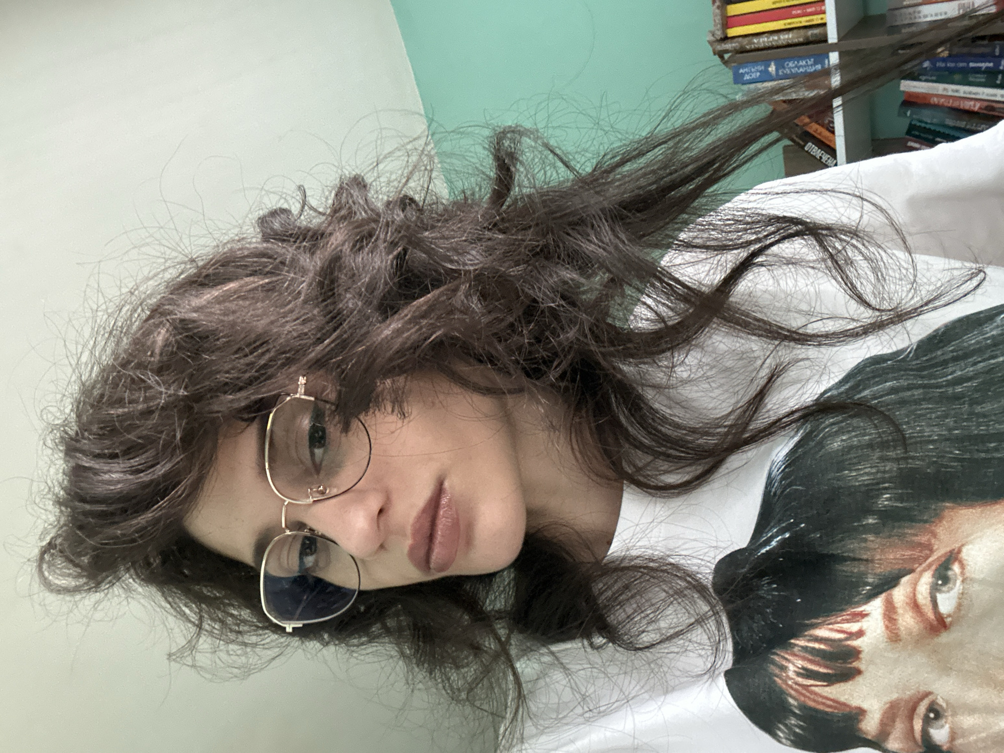 This media file shows a close-up, rotated selfie of a young woman. She has long, dark, slightly messy hair that frames her face and extends across the image. She is wearing round, gold-rimmed glasses with blue-tinted lenses. Her lips are slightly parted and appear to have a nude or light pink lipstick on. The woman is wearing a white t-shirt with a graphic print of a person's face on it, partially visible on the right side of the frame.
The background includes a light turquoise wall and a bookshelf filled with books. The books have visible spines with text, and some appear to be in Cyrillic script. The overall lighting suggests it is daytime indoors. The scene is casual and personal, likely taken in a home environment. The woman's expression is neutral, with a slight hint of a pout. The image appears to be a self-portrait, captured from a low angle.