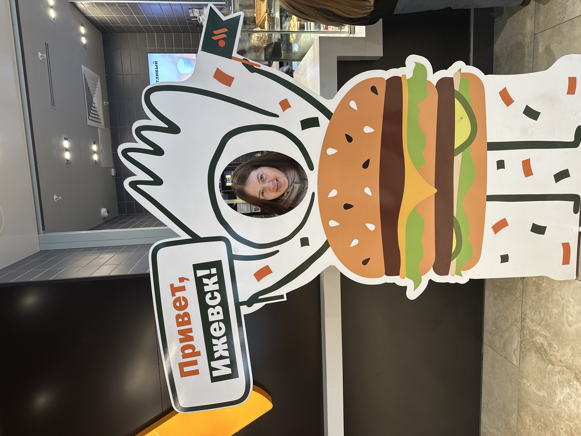 A woman is smiling and looking through a hole in a cardboard cutout of a cartoonish burger character, positioned inside a building. The cutout also features a sign that reads "Привет, Ижевск!" which translates to "Hello, Izhevsk!" in Russian, indicating the location is in Izhevsk, Russia. The background shows an interior with a food display case and other customers nearby. The environment appears to be a fast-food restaurant or cafe setting.