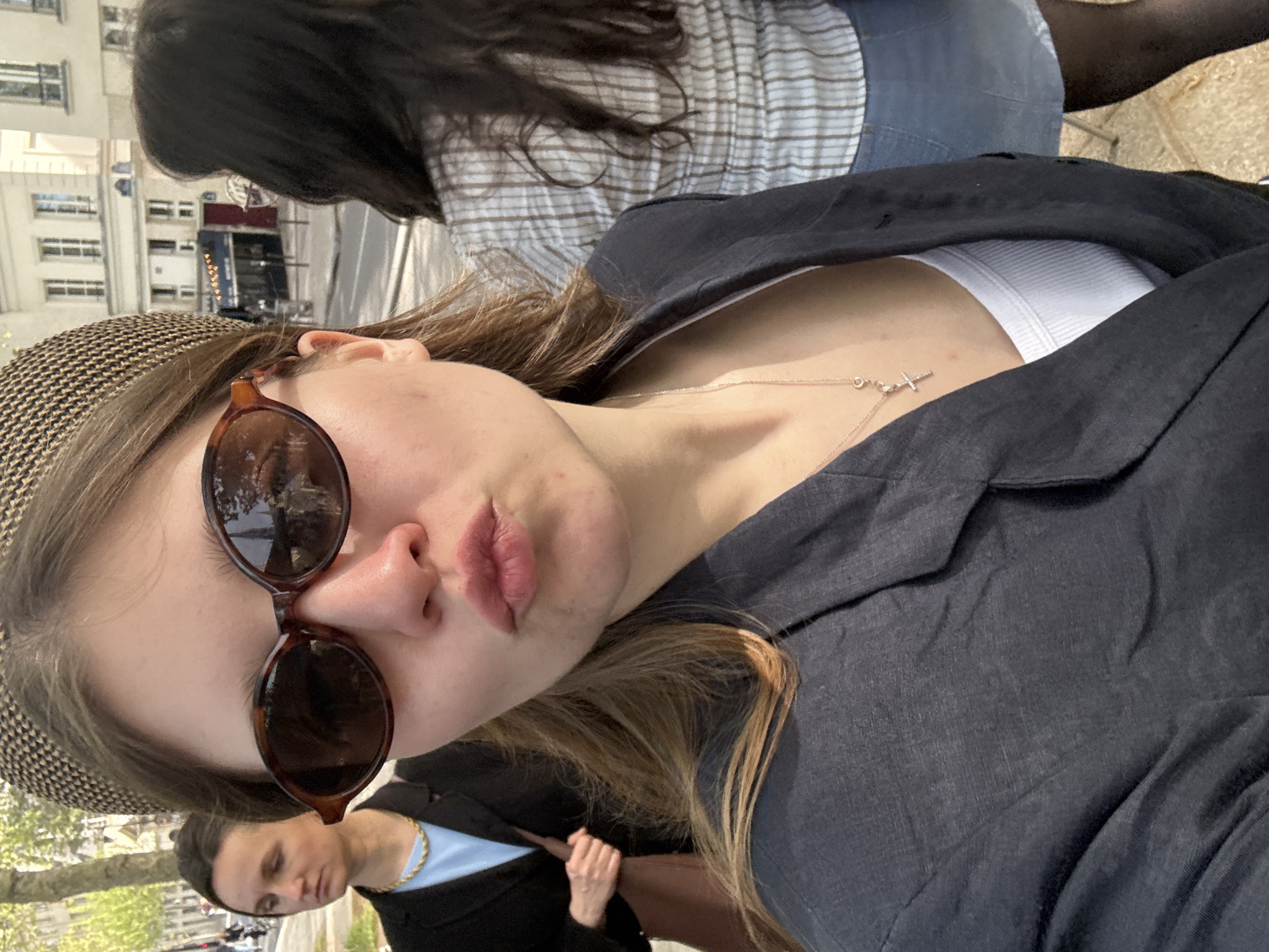 A close-up, eye-level shot shows a woman in Paris, France. She wears a patterned hat and round sunglasses. Her light brown hair is loose. A silver necklace with a cross pendant rests on her chest, visible above a white undershirt. She wears a black jacket. Behind her and to the left, another individual wears a black jacket and a blue shirt with a gold chain. Further back, buildings line a street, with trees in the foreground. Another person sits behind her and to the right and wears a black and white striped shirt.