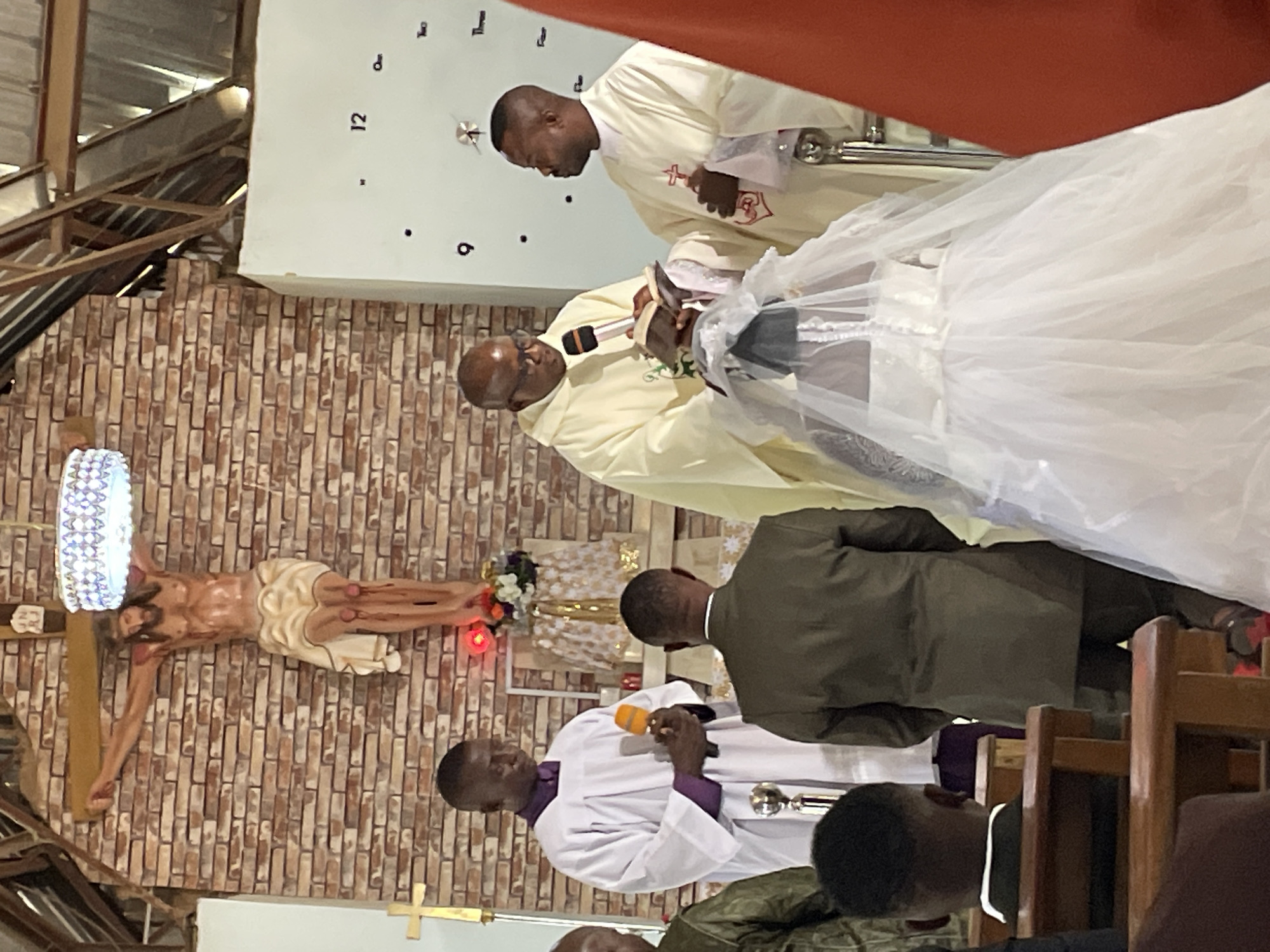 The image shows a wedding ceremony inside a church in Jos, Nigeria. A bride in a white wedding dress and veil stands facing a priest who is speaking into a microphone. Another priest stands to the bride's left. A man in a green suit stands behind the bride. A crucifix hangs on the brick wall to the left of the altar. A clock is partially visible on the wall above the crucifix. Other church members and guests are seated in the foreground.