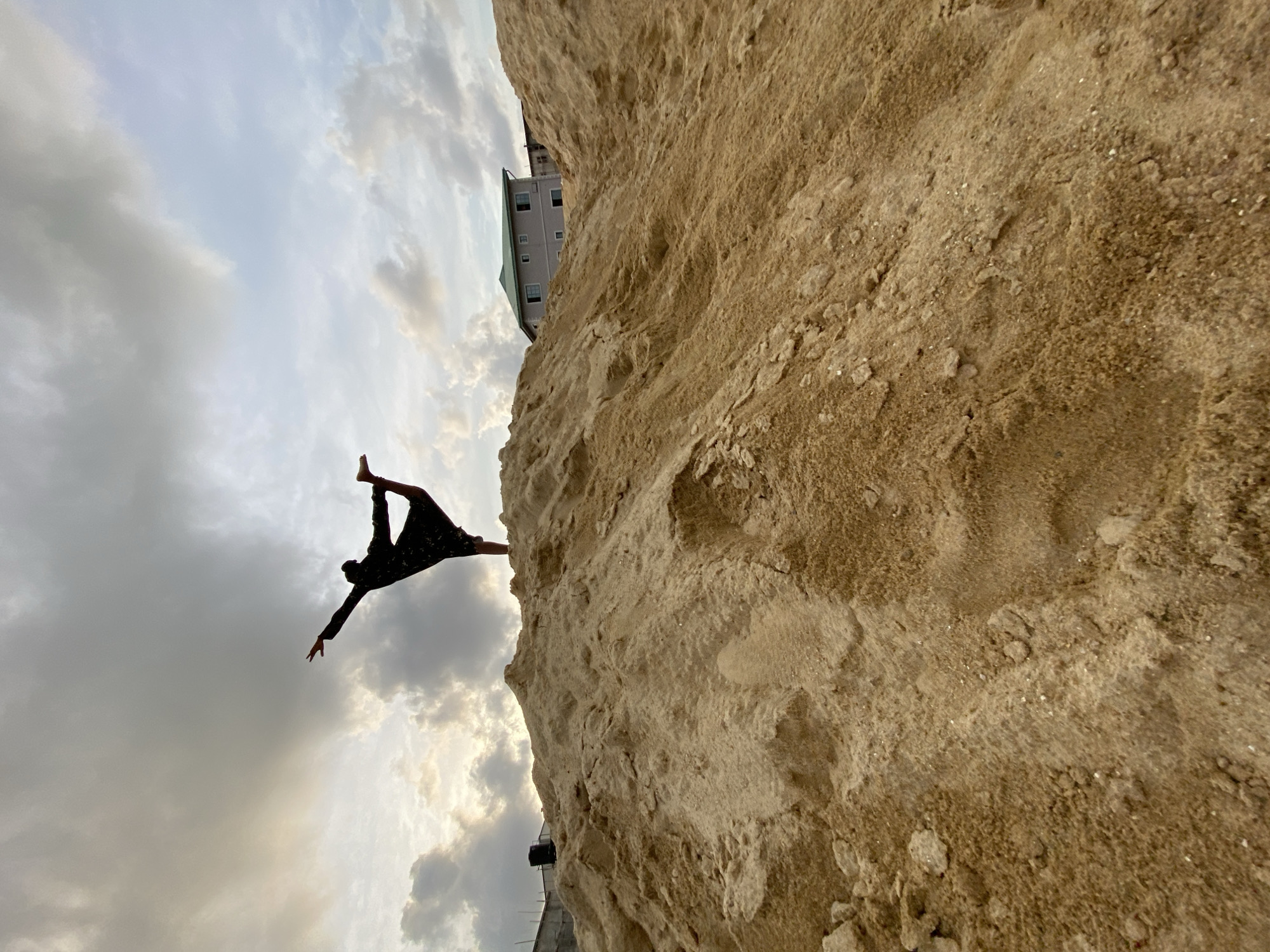 An outdoor scene in Iranla, Nigeria, depicts a silhouetted individual performing a yoga-like dancer's pose atop a large, uneven mound of light brown sand. The person is balanced on their right leg, with their left leg bent backward and upward, grasped by their left hand, while their right arm is extended forward and slightly upward. They are wearing a dark, possibly patterned, long-sleeved garment. The sky above is heavily clouded, with patches of bright light visible through breaks in the clouds. In the background, partially obscured by the sand pile, a two-story residential building with light-colored walls and a green sloped roof is visible to the right. To the far left, a darker, possibly unfinished concrete structure with a black water tank on its roof is faintly discernible behind the sand.