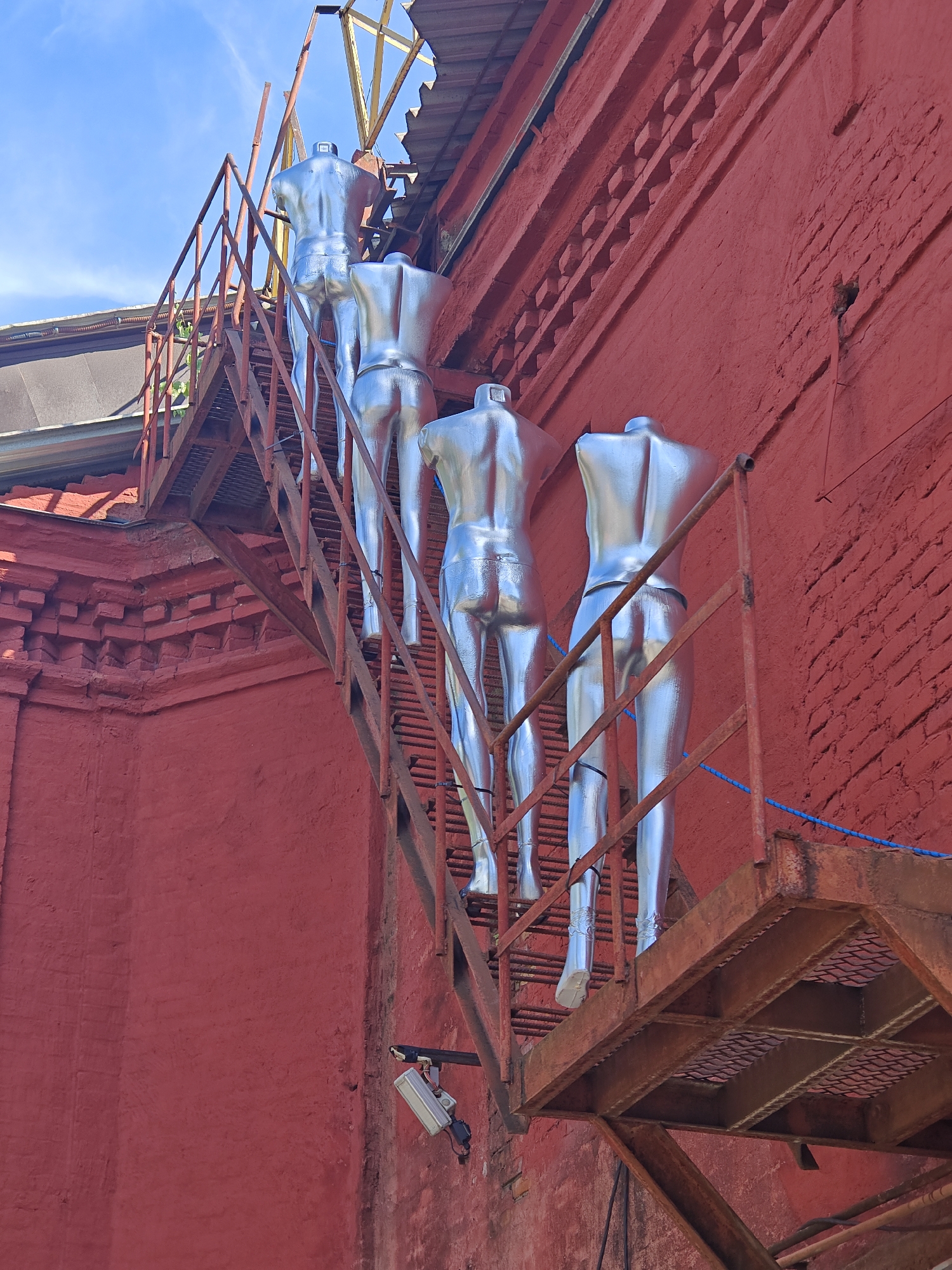 A metallic silver mannequin stands on a rusty metal fire escape staircase attached to a bright red brick building. The mannequin is the lower half of a human figure, visible from the waist down, with metallic leggings and feet. The fire escape is old and shows signs of rust and wear. The building's red brick wall is textured and has a decorative brick trim near the roofline. Above the building, a blue sky with white clouds is visible. A security camera is mounted on the red wall below the fire escape. The overall scene suggests an outdoor urban environment in Moscow, Russia.