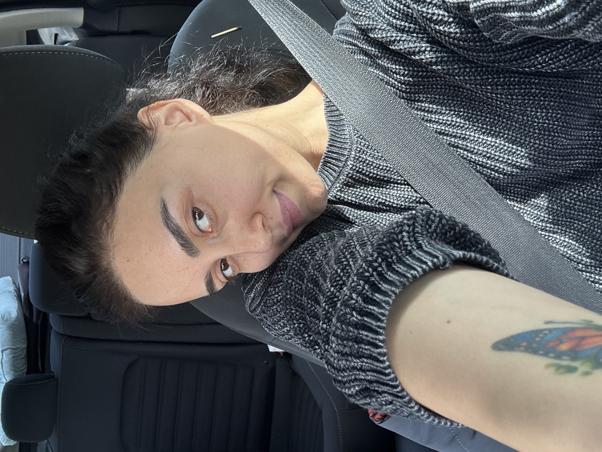 A person with dark hair is wearing a grey and white patterned sweater and a seatbelt in what appears to be a car. The person has a tattoo on their arm. The interior of the car is black. A small, thin stick-like object is visible near the top of the headrest. The location is not discernible from the image.