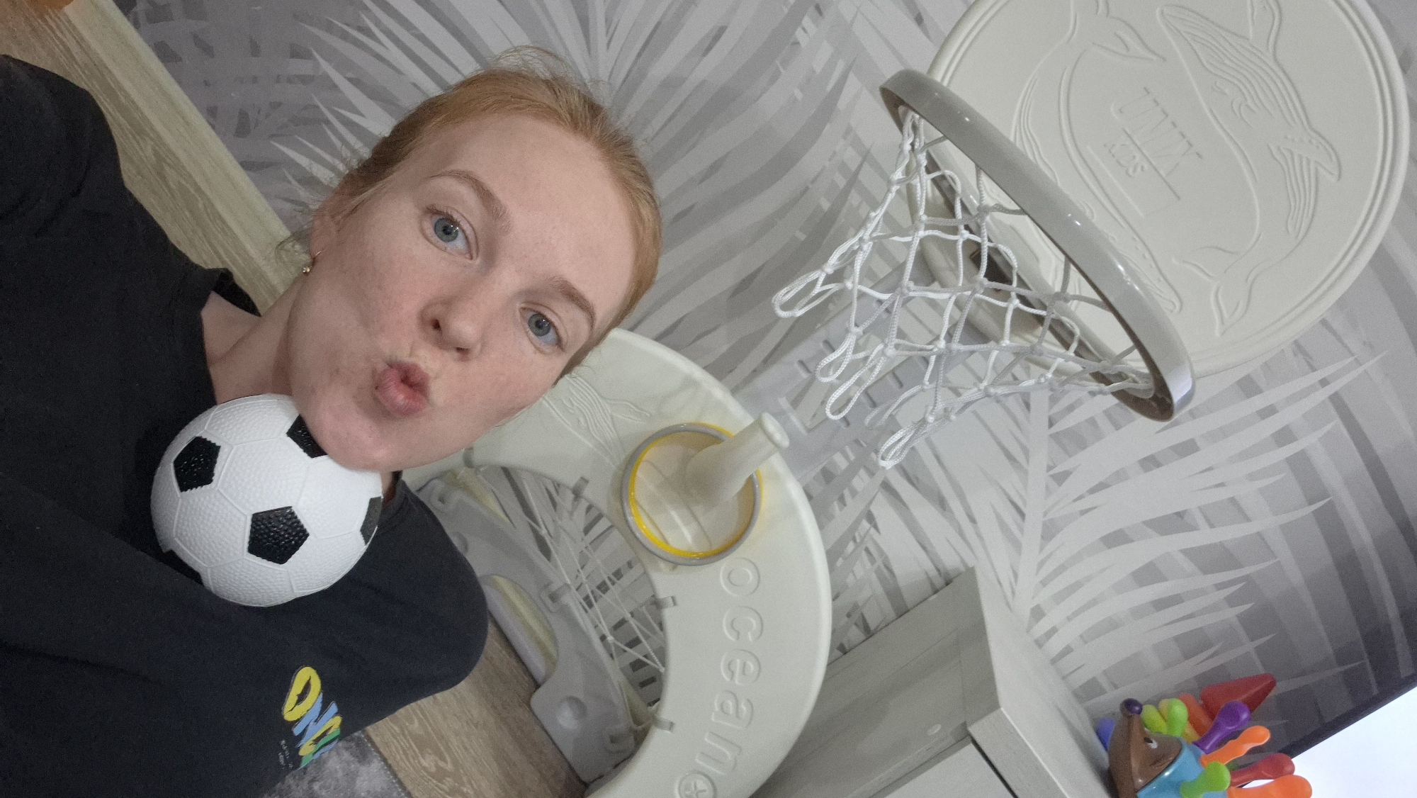 The image shows a fair-skinned woman with red hair and blue eyes holding a small soccer ball near her chest. She is pouting her lips and looking directly at the camera. She is wearing a black sweatshirt. Behind her is a child's basketball hoop and stand with a net. The basketball hoop's backboard has a whale design. Below the hoop, there are children's toys with brightly colored spikes. The background is a wall with a gray and white pattern. The location is in an indoor setting in the city of Chaikovsky, Russia.