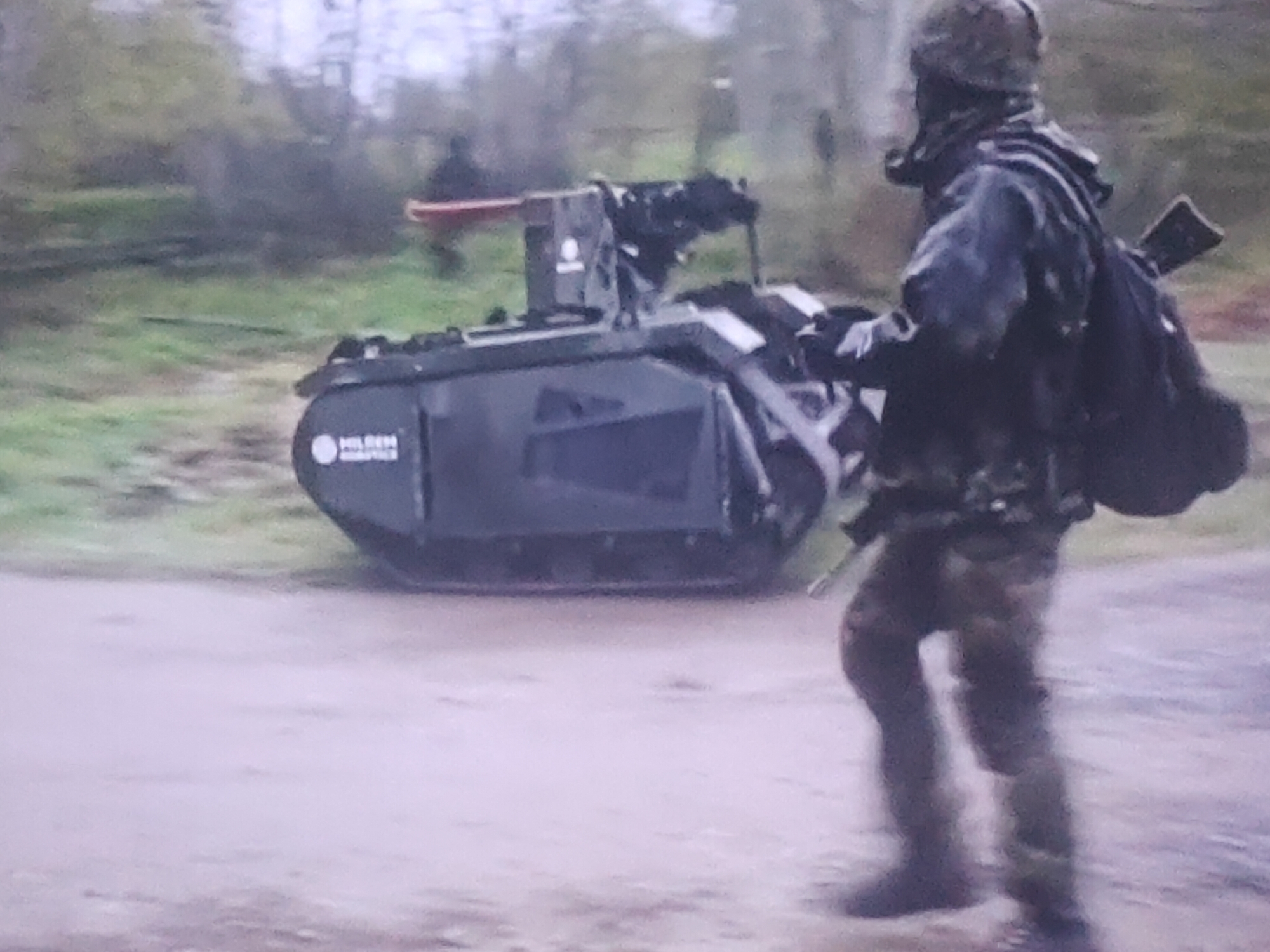 In a possibly urban or suburban outdoor setting, a person dressed in camouflage attire, including a helmet and what appears to be a gas mask, is captured in motion. They are carrying a rifle slung over their shoulder and a backpack. In front of this individual is a dark-colored, tracked robotic vehicle. The robot is equipped with a weapon system mounted on top. The background features vegetation and trees, suggesting a natural or semi-natural environment. The ground is a light-colored, possibly unpaved surface. The image is pixelated and somewhat blurry, making precise identification of certain details challenging.