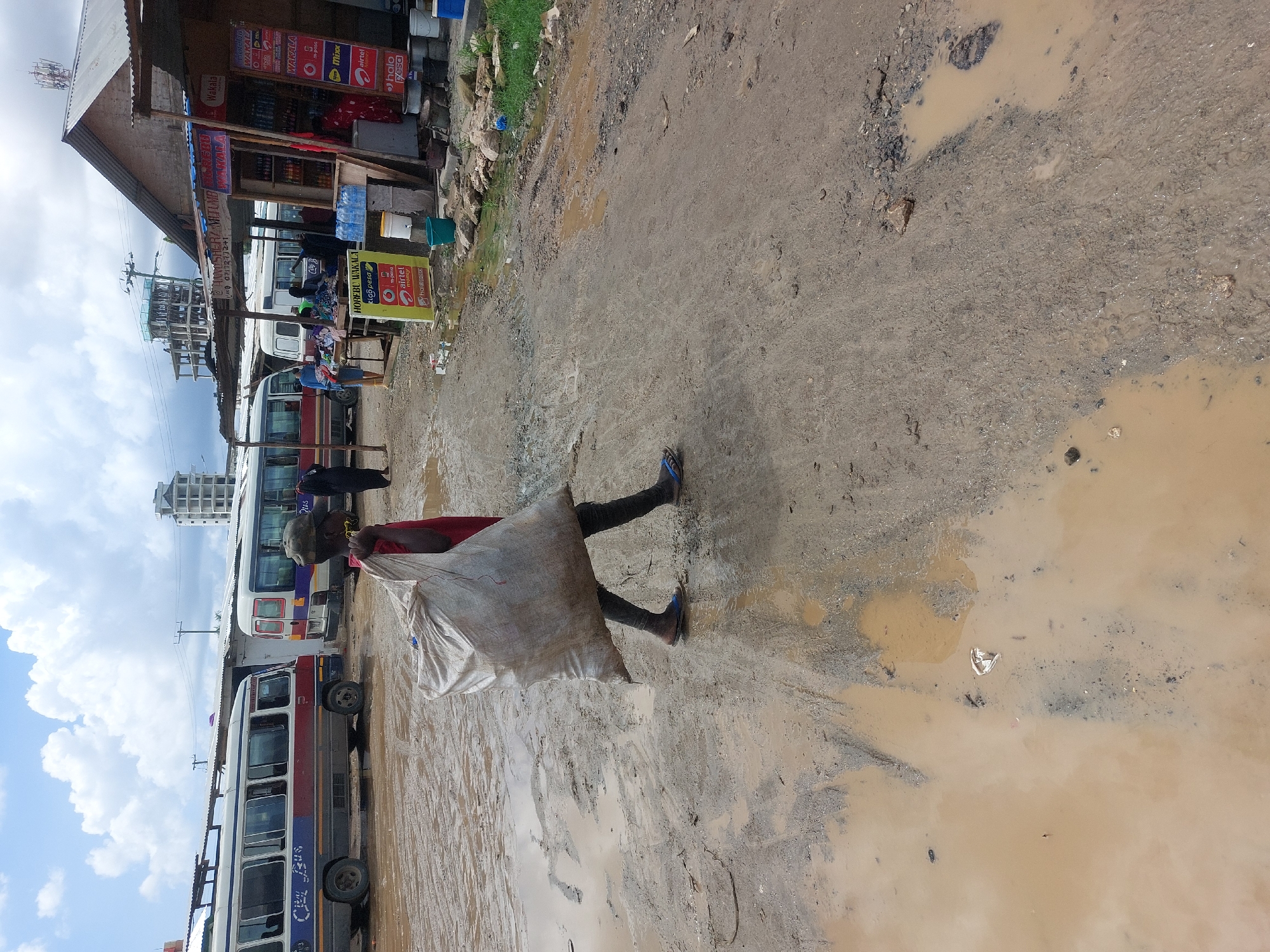 The image captures a bustling, muddy street scene in what appears to be an urban or peri-urban area, consistent with Dar es-Salaam, Tanzania. The foreground is dominated by a person walking left-to-right across the frame, carrying a large, heavy-looking white sack over their left shoulder. They wear dark trousers and blue sandals, navigating through significant puddles and uneven, dirt terrain.
To the left, a row of buses, likely local "dala-dalas," are parked or queuing. One bus is distinct with red and white colors. Behind the buses, a simple, open-fronted shop or stall with a corrugated metal roof is visible. The ground is heavily rutted, muddy, and dotted with large, murky water puddles, suggesting recent rainfall or poor drainage.
Notable details include several advertisements on the shop and on freestanding signs. Visible text includes "Wakala" (agent), "Horebu Wakala," and mobile money service brands such as "M-Pesa," "airtel money," and "halo pesa." Other signs show "Mixxx" and a partial phone number "071727." Various goods are displayed in and around the shop, including bottled drinks and buckets. In the background, under a partly cloudy sky, there are communication towers and what appears to be an unfinished building. The scene depicts everyday life and commerce in a challenging, muddy environment.