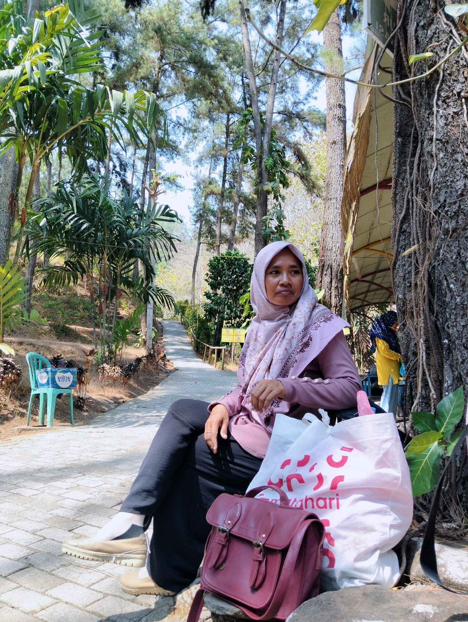 A woman wearing a light purple hijab and a matching long-sleeved shirt sits on a bench in a park. She is wearing black pants and light brown slip-on shoes with white socks. Next to her are two shopping bags, one large white bag with red lettering that reads "Matahari," and a smaller maroon satchel. In the background, a paved path winds through a wooded area with palm trees and tall, thin trees. To the left of the path, a bright blue plastic chair sits next to a crate of bottled water. In the far background, another woman in a yellow shirt and blue hijab is visible, along with a structure with a brown roof. The scene appears to be outdoors in a tropical or subtropical climate, given the presence of palm trees and the bright sunlight.