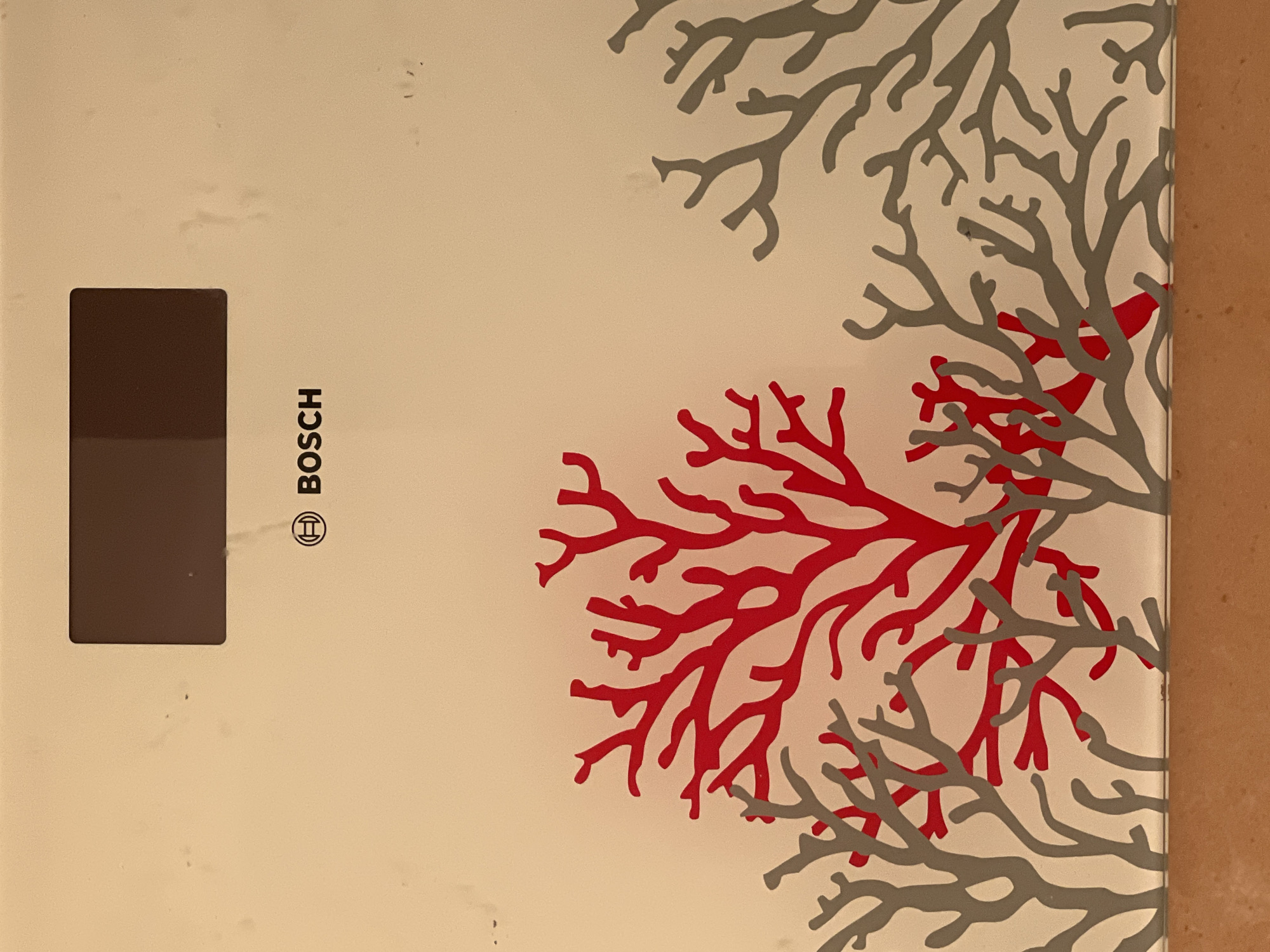 The image shows a close-up of a beige-colored scale. At the top center of the scale is a dark rectangular display. Below the display is the Bosch logo. The lower portion of the scale features a decorative pattern of red and grey coral-like branches. The background is a solid beige color with some subtle smudges and speckles. The bottom edge of the scale is visible, with a warm brown surface beneath it. The location in the city of Minsk, Belarus is not discernible from the image.