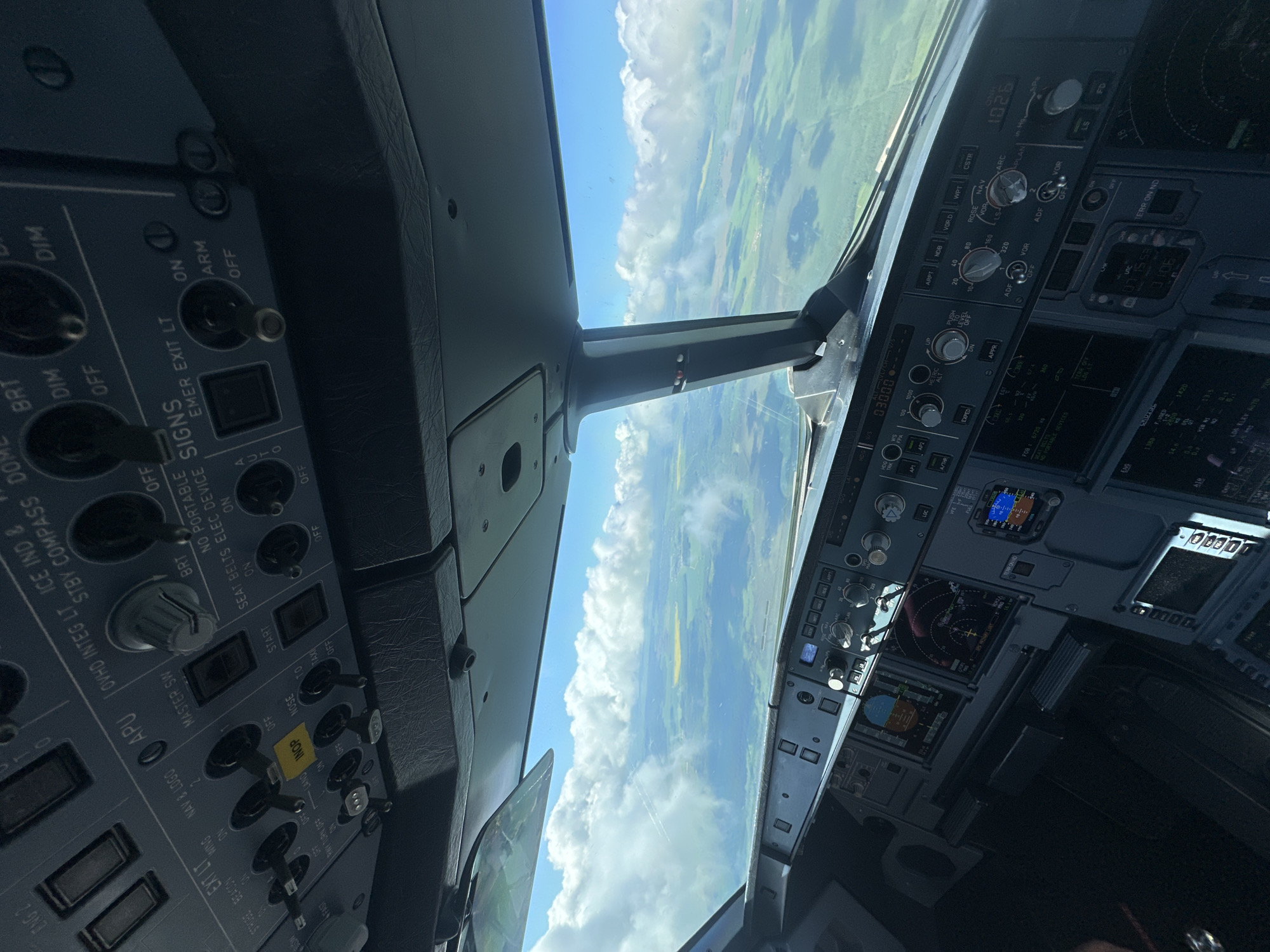 The image shows the interior of an aircraft cockpit during flight. Through the windshield, a landscape of green fields and white clouds under a blue sky is visible. The control panels on both sides of the cockpit contain various switches, knobs, and displays. These panels are labeled with terms like "MASTER SW", "STROBE BEACON", "SEAT BELTS", "EMER EXIT LT", "VOR", "NAV", and "FD". The heading display shows 03000, along with other various readings on other systems. There are no people visible in the shot, only the pilot environment. There is no indication this is in Moscow.