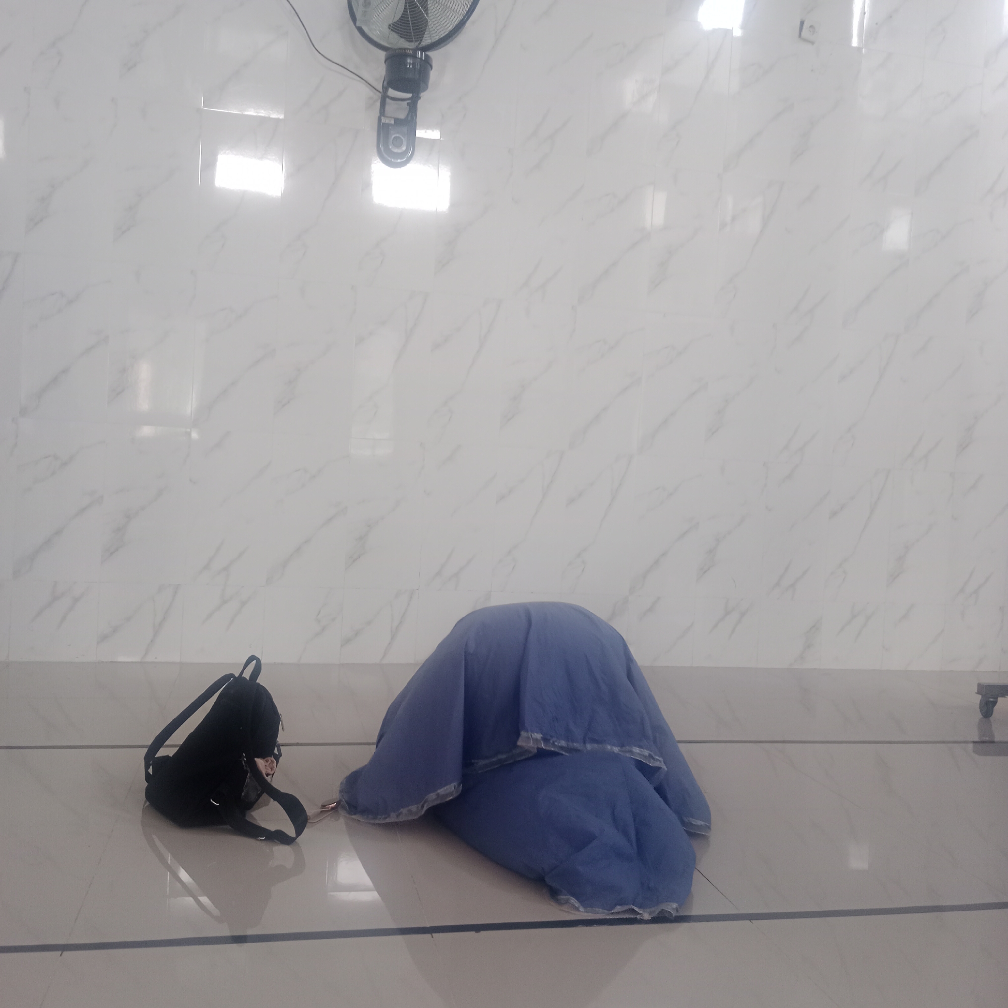 In Karanganyung, Indonesia, an indoor scene is depicted with a white marble-patterned wall and a tiled floor. A black backpack rests on the floor to the left of the frame. To the right, a person is covered by a blue garment, appearing to be lying down, with a hint of their feet visible at the bottom right edge of the frame. A ceiling fan is mounted high on the wall in the upper portion of the image. The lighting suggests the presence of overhead lights reflecting off the wall and floor.