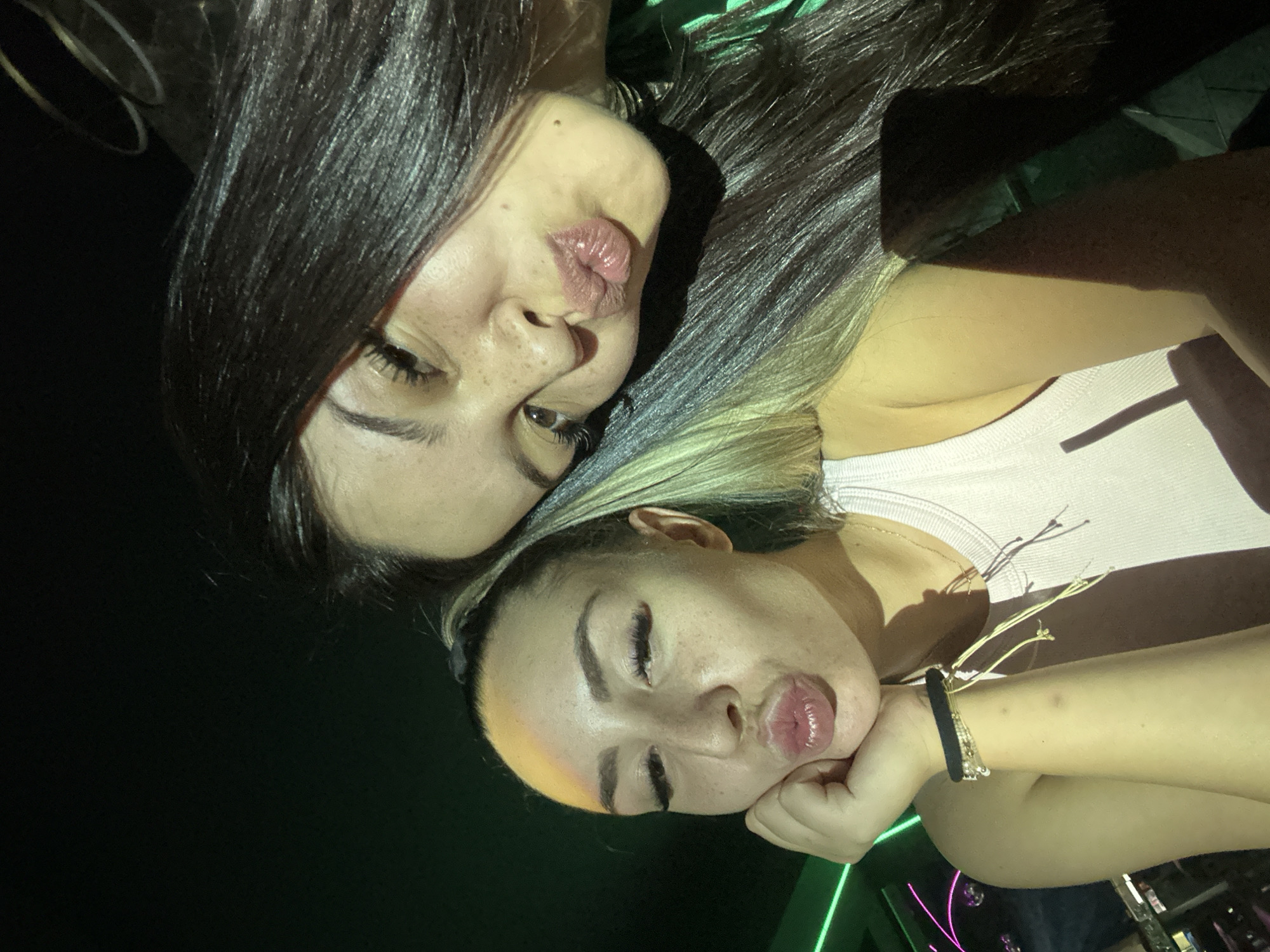 The image is a selfie taken by two young women in what appears to be a dimly lit nightclub or bar. The woman in the foreground, on the left, has blonde-streaked hair pulled back in a ponytail, and is resting her chin on her hand. She is puckering her lips and wearing a light pink tank top and several bracelets on her left arm. The woman in the background, on the right, has dark hair and is leaning her head towards the woman in the foreground, also puckering her lips. Her face is partially obscured by her hair and the angle of the selfie. The background is dark with some neon green lighting visible on the left side, suggesting a nightlife setting. There is no visible indication of the specific location within the city of Sliven, Bulgaria.