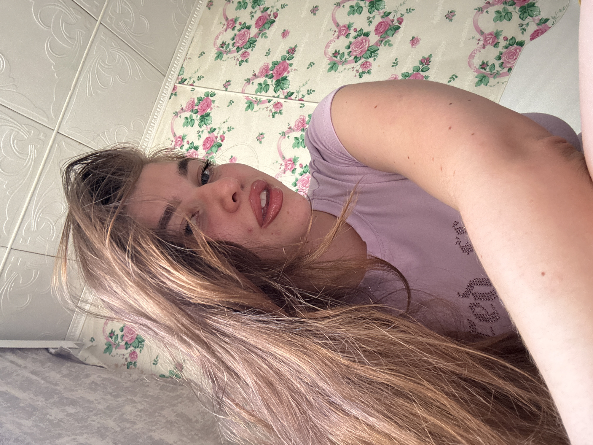 The image shows a close-up, low-angle selfie of a young woman with long, wavy brown hair. She is wearing a light purple t-shirt with a sparkling design on the chest. Her lips are glossy and her eyes are looking towards the camera. The background consists of a wall covered in floral wallpaper with pink roses and green leaves, and a textured ceiling above. The setting appears to be indoors, likely a bedroom. There is no information available to determine the specific location within Мещёры, Russia.