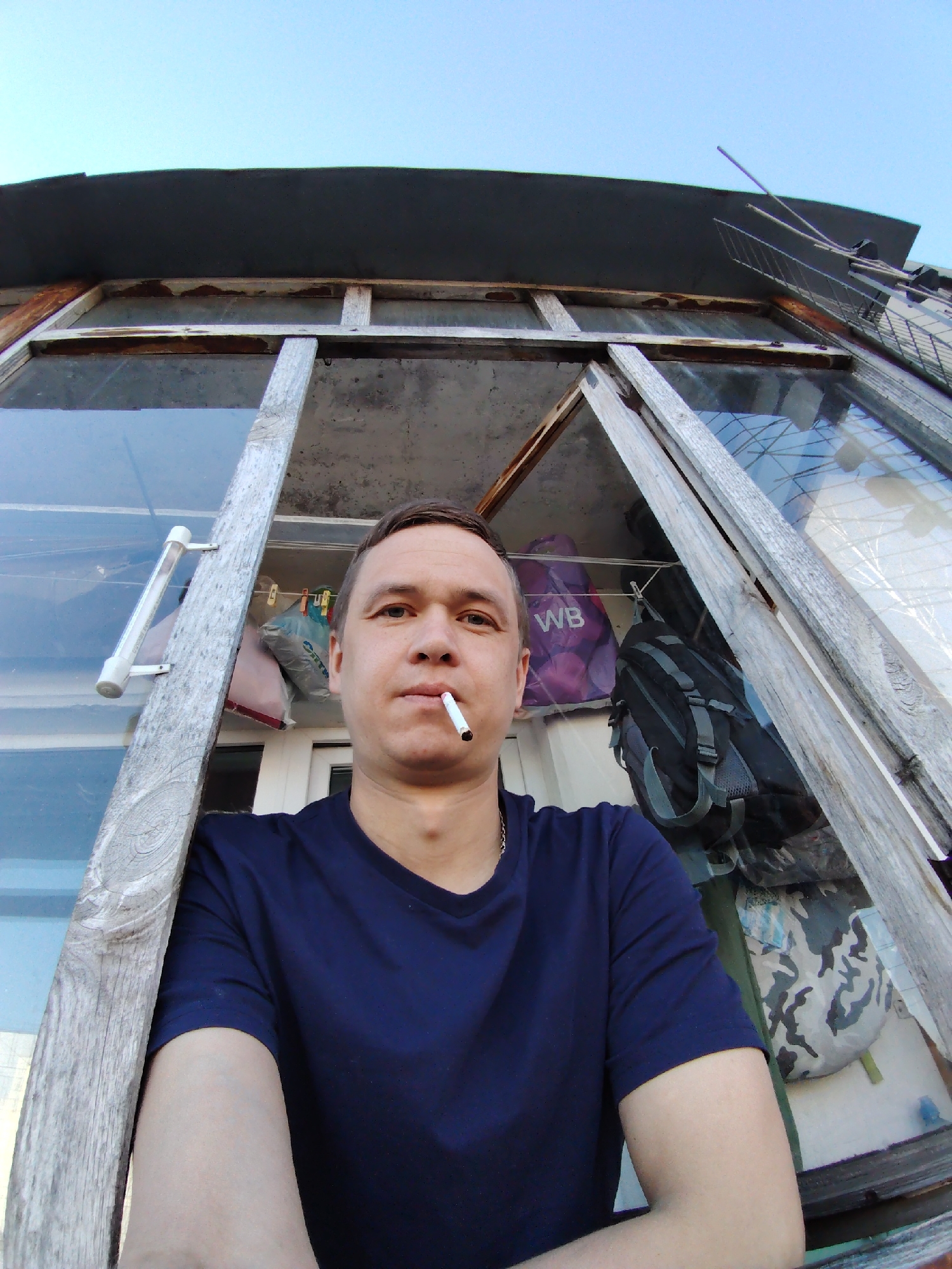 A man stands in an open, weathered window frame of an enclosed balcony. He holds a cigarette in his mouth. The balcony is cluttered with items, including bags, backpacks, and clothes hanging on a line. Above the balcony is a dark roof and an antenna. The photograph was taken at an upward angle, possibly with a wide-angle lens. The location is Chaikovsky, Russia.