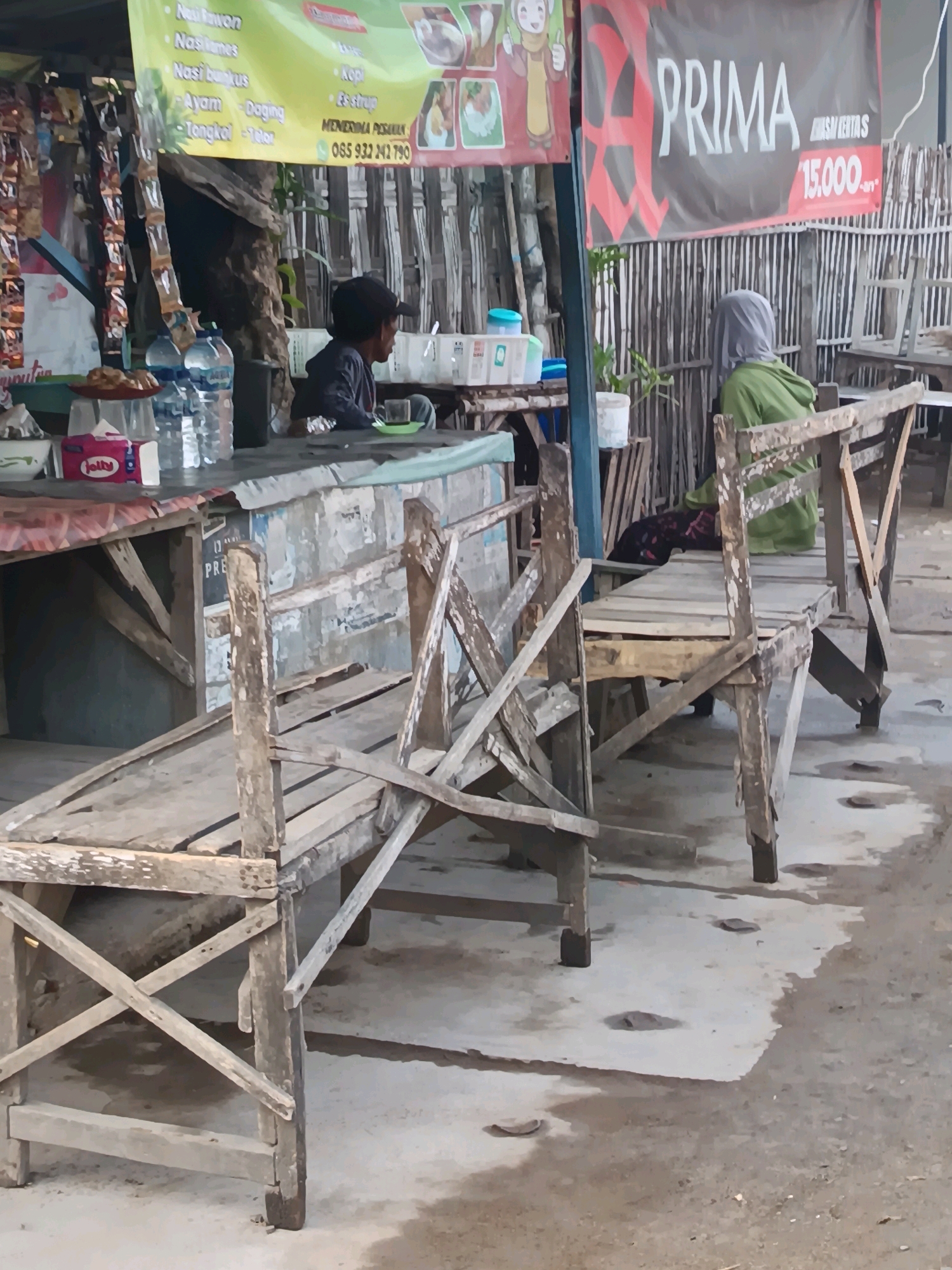 The image captures a street-side food stall in Blega, Indonesia. In the foreground, two weathered wooden benches are positioned on a concrete ground, one angled towards the left and the other towards the right. Behind the benches, a makeshift stall features a counter laden with various items, including bottles of water, a box of tissues, and some food. Two individuals are present: a person wearing a dark cap is seated at the counter, and another person wearing a green hoodie and a headscarf is seated on the bench to the right, facing away from the camera. A large banner advertising "PRIMA" with a price of "15.000" is displayed prominently on a fence behind the stall. The environment suggests a casual, outdoor dining setting.