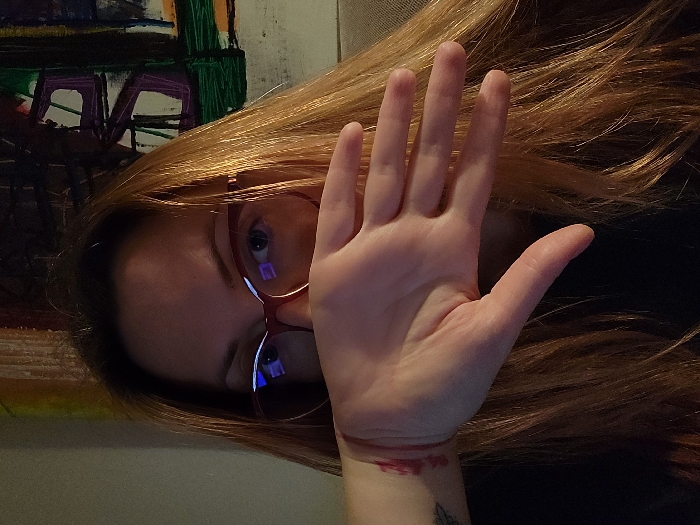 A person with reddish-blonde hair and red-framed glasses is looking downward, their gaze visible through the glasses. A hand is raised, fingers spread, obscuring part of their face. A tattoo is visible on the wrist, near what appears to be a wound or red mark. The background includes an abstract painting with colors like green, purple, and yellow. The image is rotated, with the subject appearing sideways.